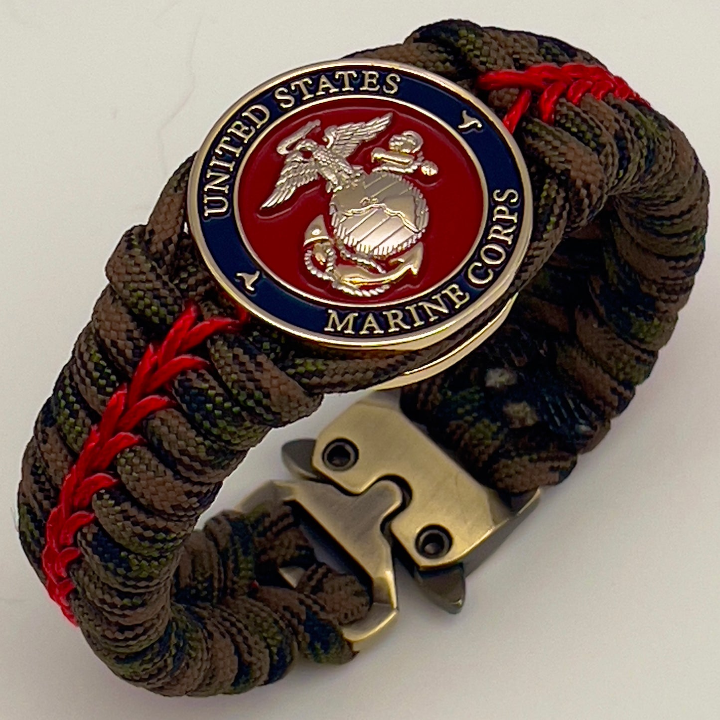 Thick, semi-rigid and comfortable braided United States Marine Corps bracelet featuring a 24K gold and silver plated USMC officer Eagle Globe and Anchor medallion, a gold Cobra style tactical buckle and woven with Woodland Camo 550 paracord and Imperial Red micro cord.