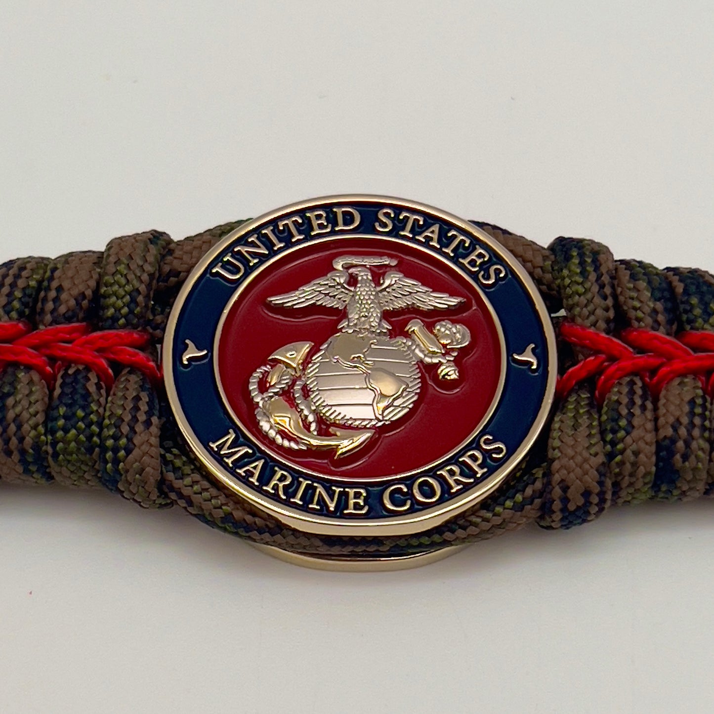Thick, semi-rigid and comfortable braided United States Marine Corps bracelet featuring a 24K gold and silver plated USMC officer Eagle Globe and Anchor medallion, a gold Cobra style tactical buckle and woven with Woodland Camo 550 paracord and Imperial Red micro cord.