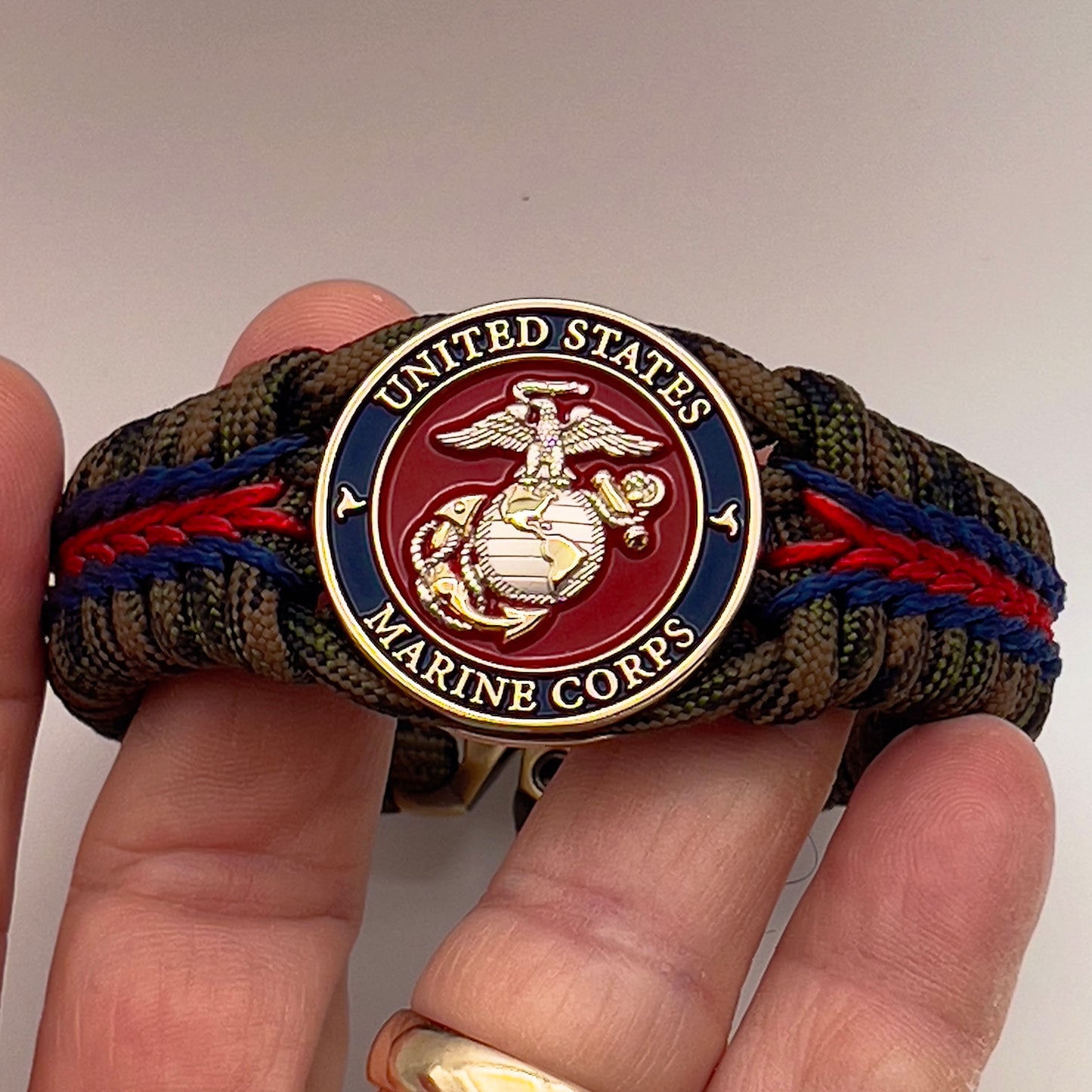 Thick, semi-rigid and comfortable braided United States Marine Corps bracelet featuring a 24K gold and silver plated USMC officer Eagle Globe and Anchor medallion, a gold Cobra style tactical buckle and woven with Woodland Camo 550 paracord and Imperial Red and Navy Blue micro cord.