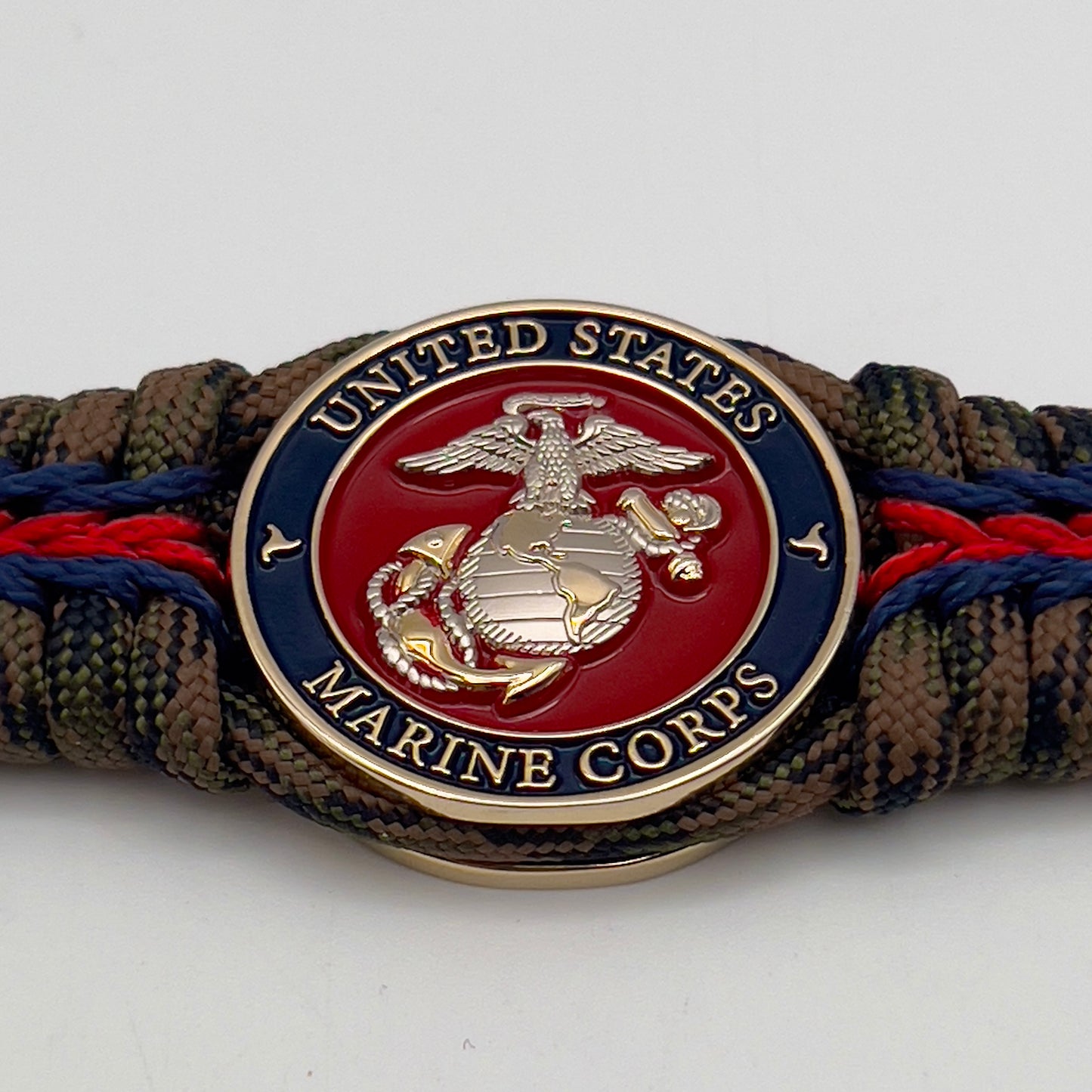 Thick, semi-rigid and comfortable braided United States Marine Corps bracelet featuring a 24K gold and silver plated USMC officer Eagle Globe and Anchor medallion, a gold Cobra style tactical buckle and woven with Woodland Camo 550 paracord and Imperial Red and Navy Blue micro cord.