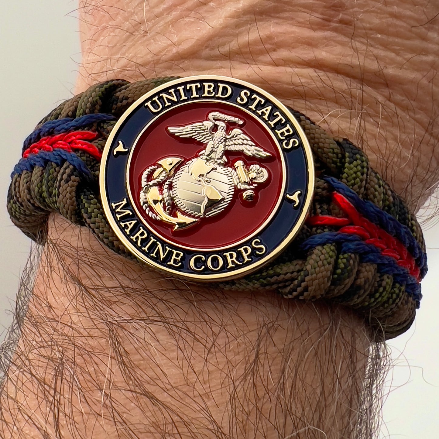 Thick, semi-rigid and comfortable braided United States Marine Corps bracelet featuring a 24K gold and silver plated USMC officer Eagle Globe and Anchor medallion, a gold Cobra style tactical buckle and woven with Woodland Camo 550 paracord and Imperial Red and Navy Blue micro cord.
