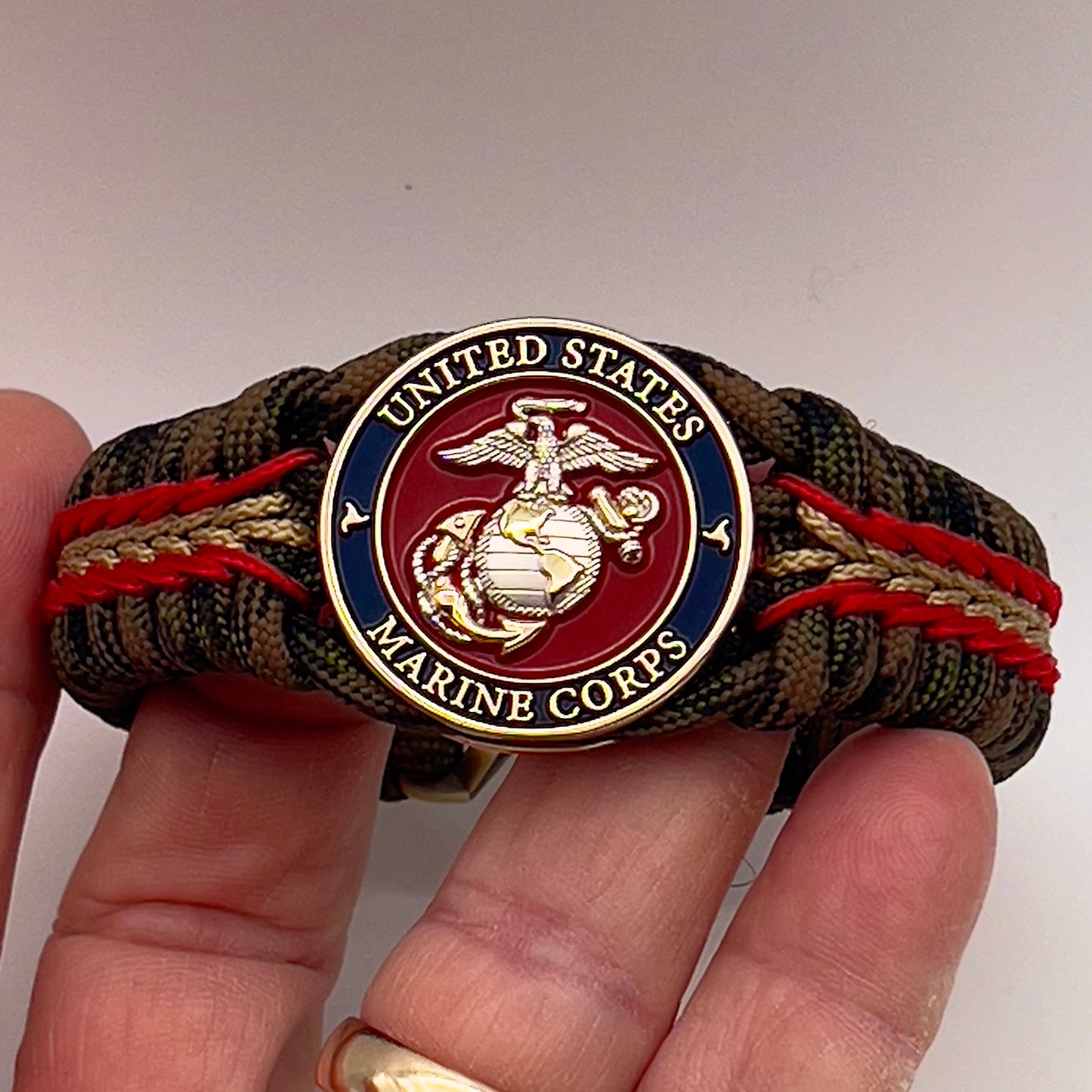 Thick, semi-rigid and comfortable braided United States Marine Corps bracelet featuring a 24K gold and silver plated USMC officer Eagle Globe and Anchor medallion, a gold Cobra style tactical buckle and woven with Woodland Camo 550 paracord and Imperial Red and gold micro cord.