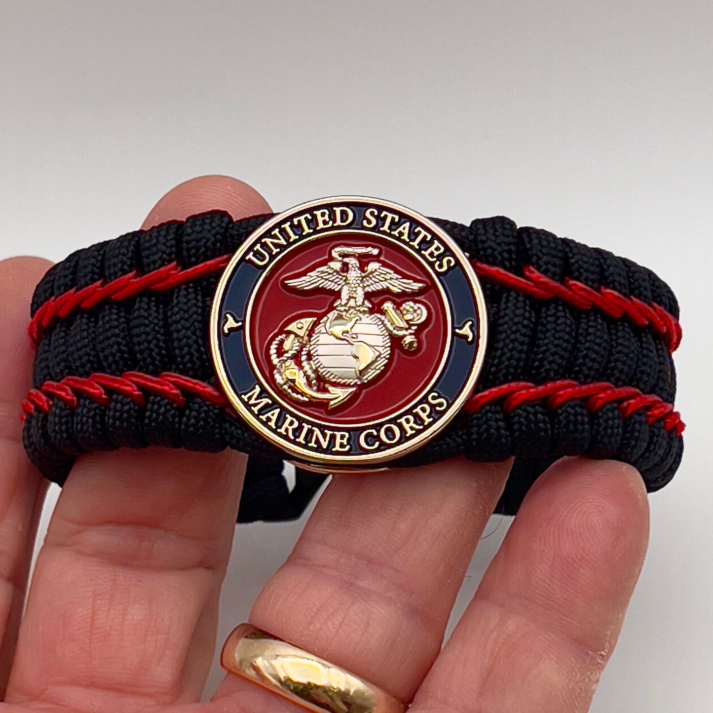Thick and comfortable braided United States Marine Corps bracelet featuring a 24K gold and silver plated USMC officer Eagle Globe and Anchor medallion, a black Cobra style tactical buckle and woven with black 550 paracord and Imperial Red micro cord.