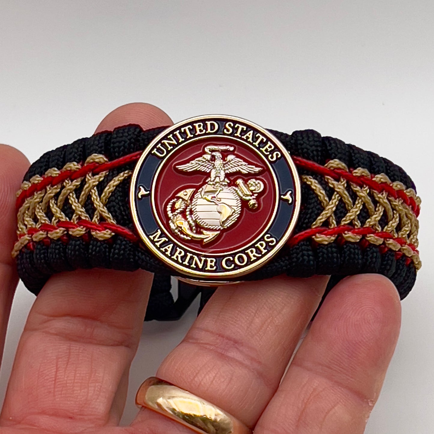 Thick and comfortable braided United States Marine Corps bracelet featuring a 24K gold and silver plated USMC officer Eagle Globe and Anchor medallion, a black Cobra style tactical buckle and woven with black 550 paracord and Imperial Red and gold micro cord.