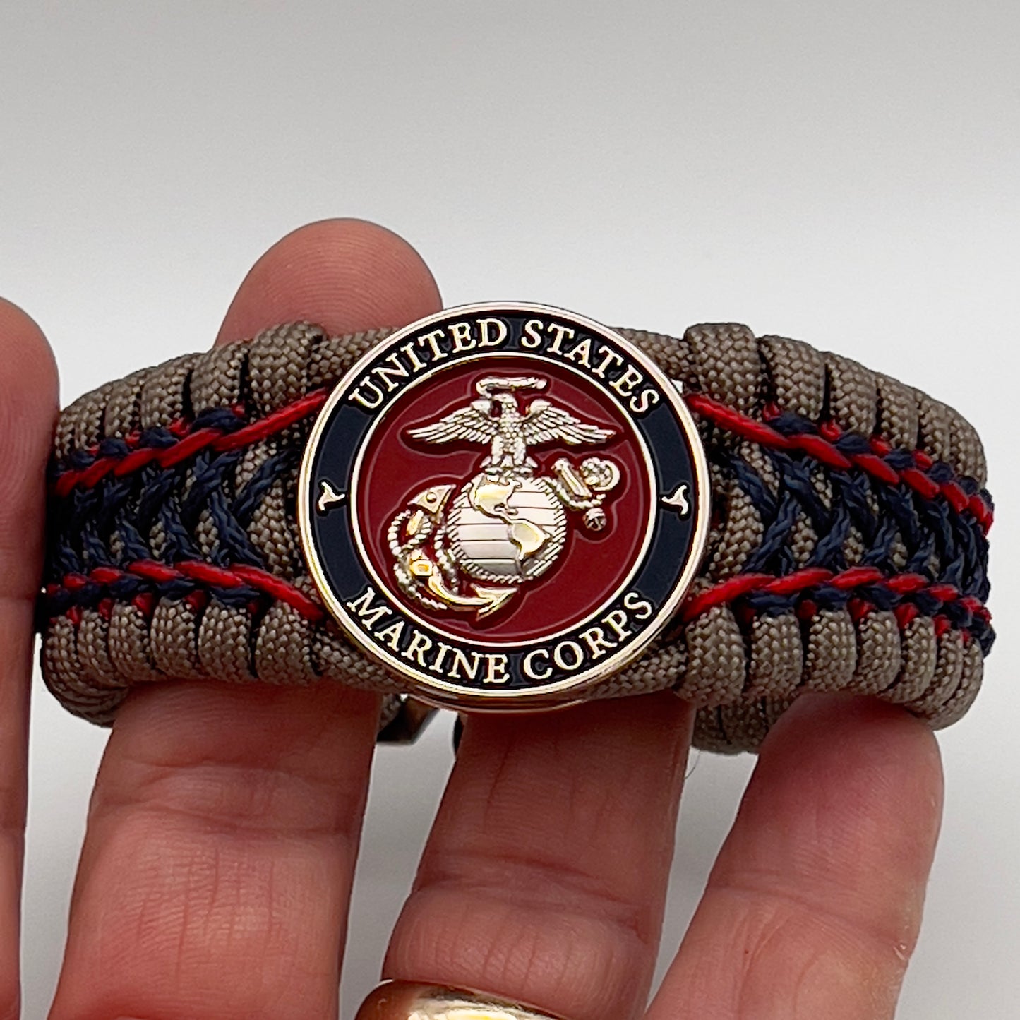 Thick, rigid and comfortable braided United States Marine Corps bracelet featuring a 24K gold and silver plated USMC officer Eagle Globe and Anchor medallion, a gold Cobra style tactical buckle and woven with tan 550 paracord and Imperial Red and Navy Blue micro cord.