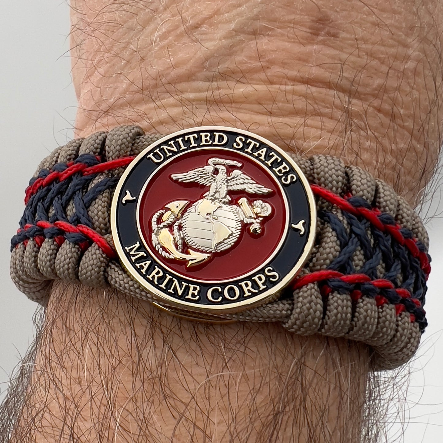 Thick, rigid and comfortable braided United States Marine Corps bracelet featuring a 24K gold and silver plated USMC officer Eagle Globe and Anchor medallion, a gold Cobra style tactical buckle and woven with tan 550 paracord and Imperial Red and Navy Blue micro cord.