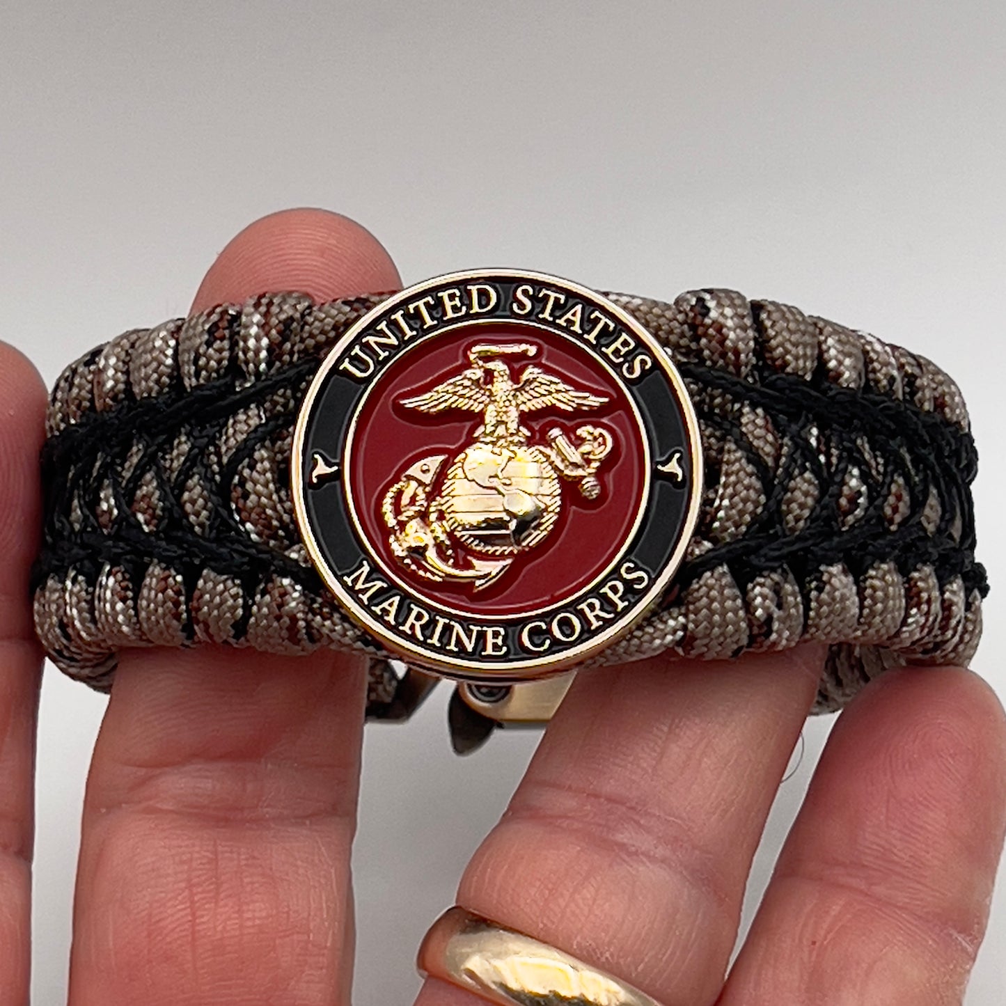 Thick, wide, rigid and comfortable braided United States Marine Corps bracelet featuring a 24K gold plated USMC Eagle Globe and Anchor medallion, a gold Cobra style tactical buckle and woven with Desert Camo 550 paracord and black nano and micro cord.