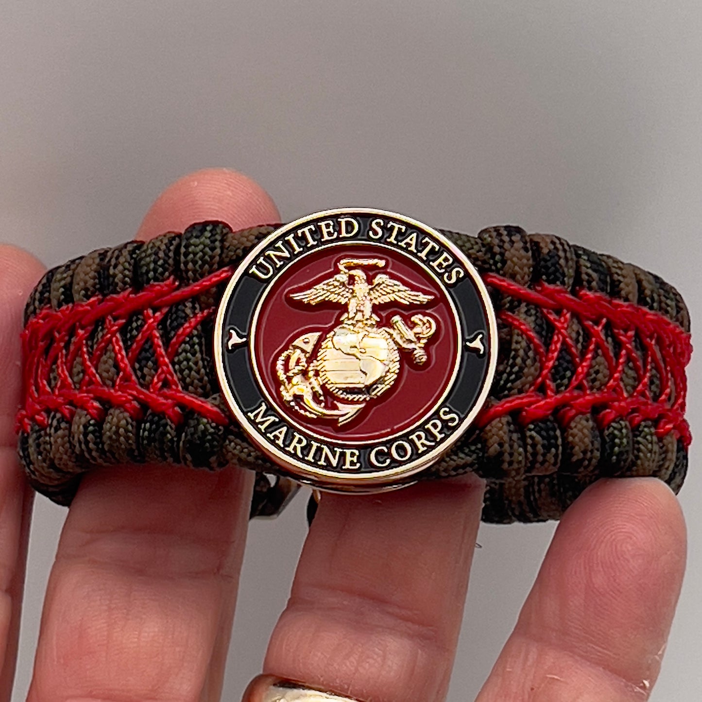 Thick, wide, rigid and comfortable braided United States Marine Corps bracelet featuring a 24K gold plated USMC Eagle Globe and Anchor medallion, a gold Cobra style tactical buckle and woven with Woodland Camo 550 paracord and Imperial Red nano and micro cord.