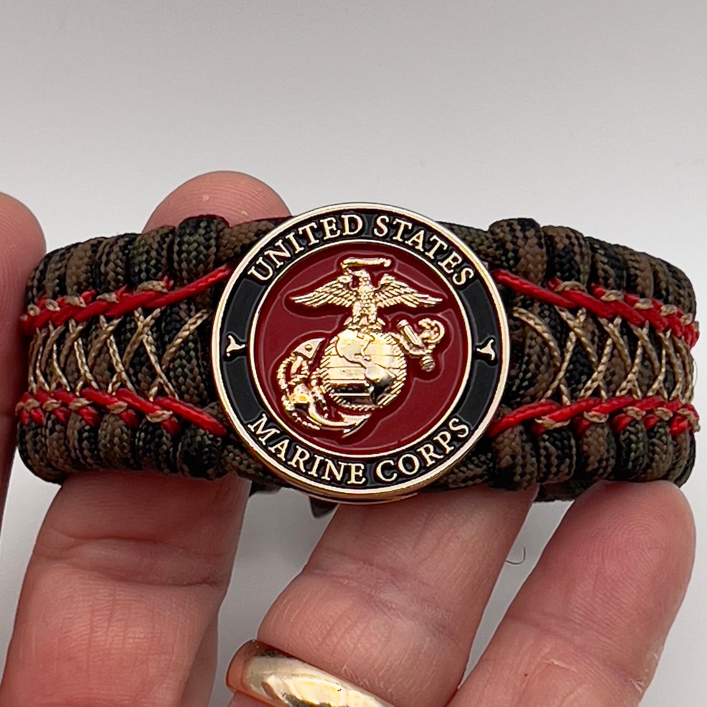 Thick, wide, rigid and comfortable braided United States Marine Corps bracelet featuring a 24K gold plated USMC Eagle Globe and Anchor medallion, a gold Cobra style tactical buckle and woven with Woodland Camo 550 paracord and Imperial Red micro and gold nano cord.