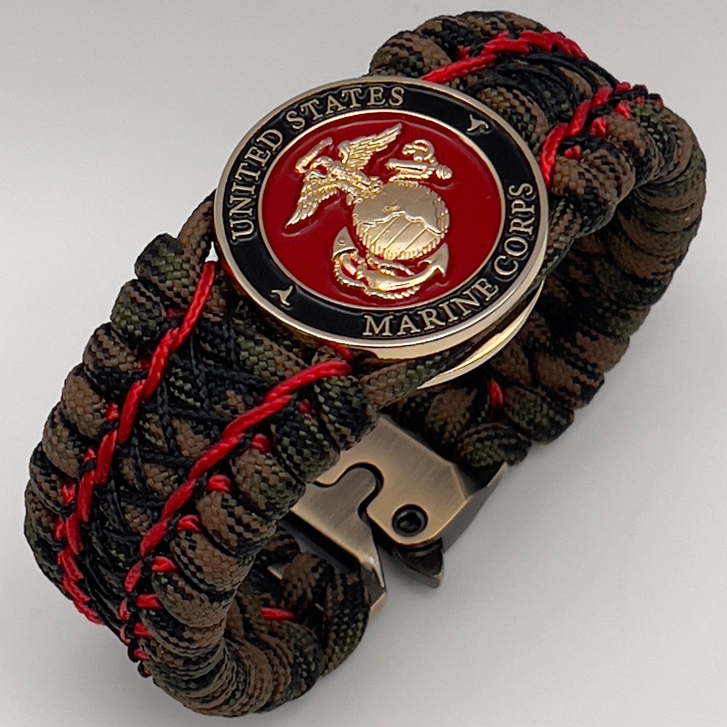 Thick, wide, rigid and comfortable braided United States Marine Corps bracelet featuring a 24K gold plated USMC Eagle Globe and Anchor medallion, a gold Cobra style tactical buckle and woven with Woodland Camo 550 paracord and Imperial Red micro and black nano cord.