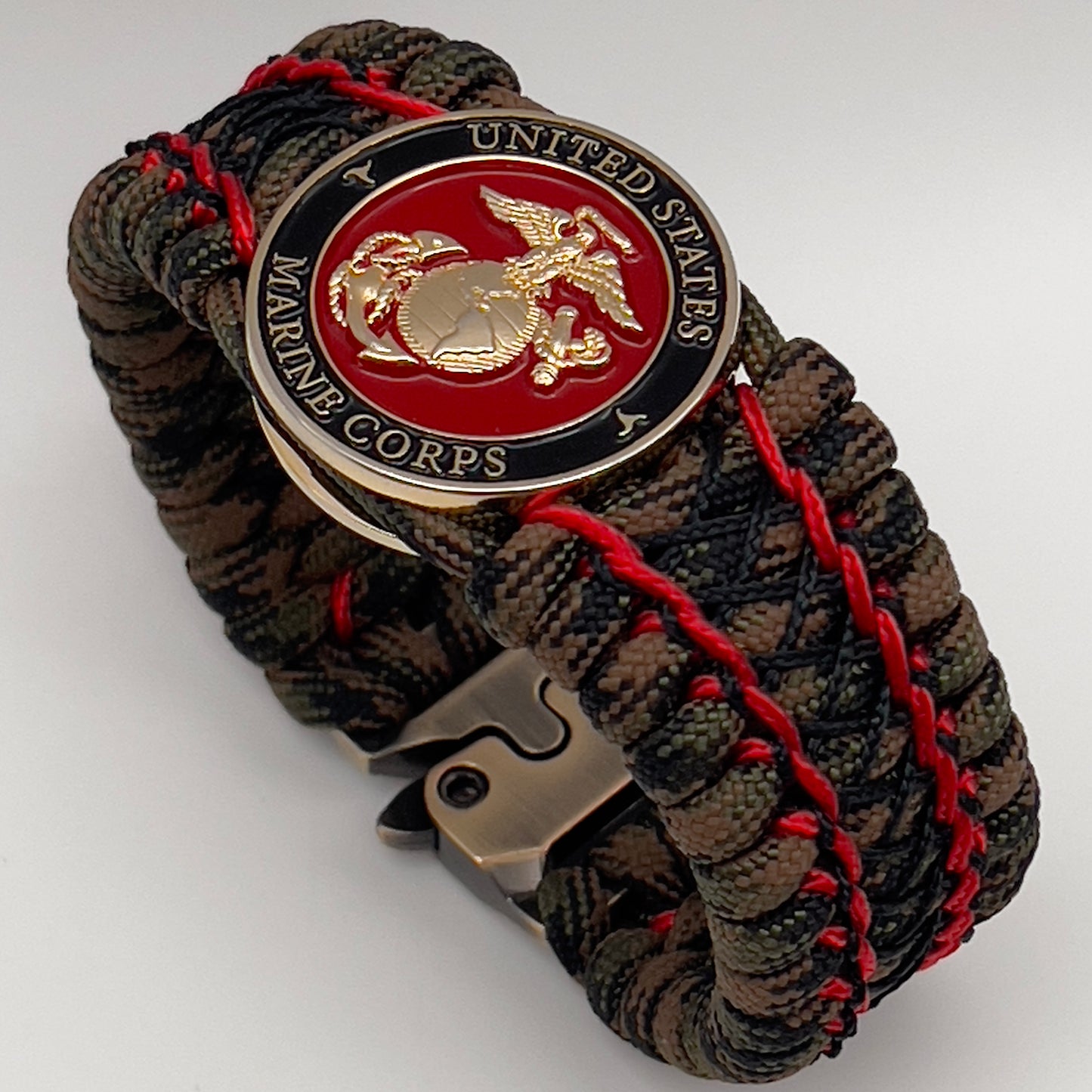 Thick, wide, rigid and comfortable braided United States Marine Corps bracelet featuring a 24K gold plated USMC Eagle Globe and Anchor medallion, a gold Cobra style tactical buckle and woven with Woodland Camo 550 paracord and Imperial Red micro and black nano cord.