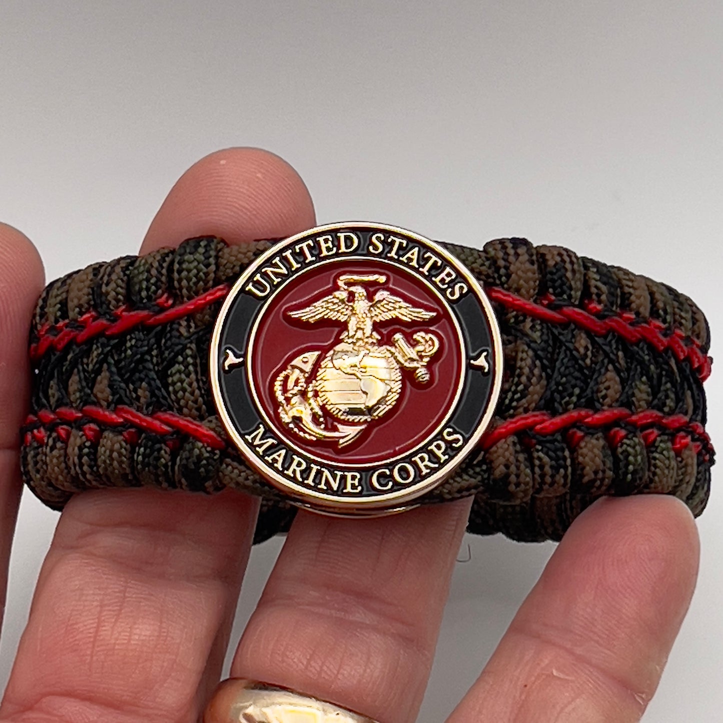 Thick, wide, rigid and comfortable braided United States Marine Corps bracelet featuring a 24K gold plated USMC Eagle Globe and Anchor medallion, a gold Cobra style tactical buckle and woven with Woodland Camo 550 paracord and Imperial Red micro and black nano cord.