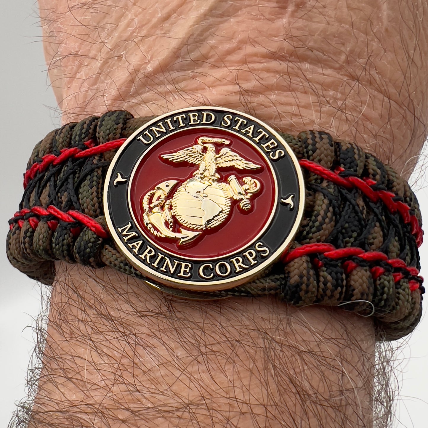Thick, wide, rigid and comfortable braided United States Marine Corps bracelet featuring a 24K gold plated USMC Eagle Globe and Anchor medallion, a gold Cobra style tactical buckle and woven with Woodland Camo 550 paracord and Imperial Red micro and black nano cord.