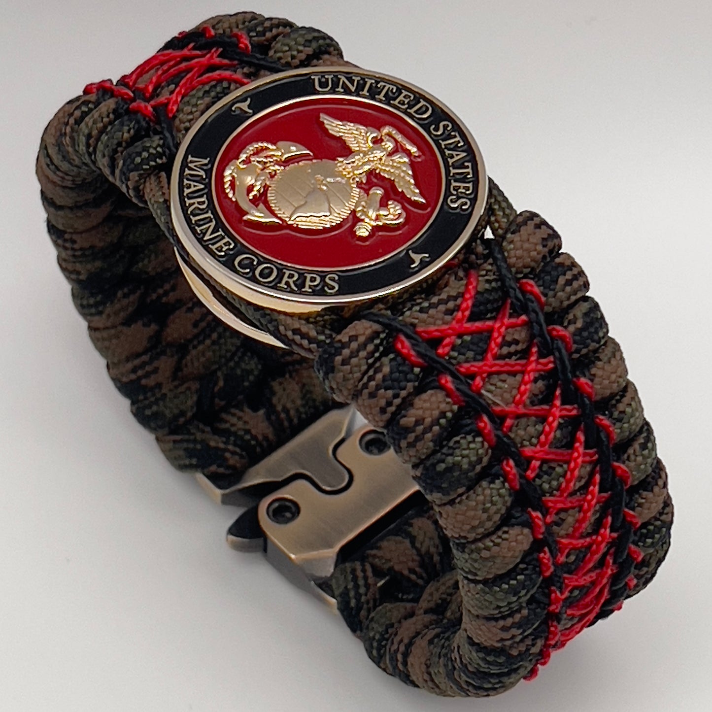 Thick, wide, rigid and comfortable braided United States Marine Corps bracelet featuring a 24K gold plated USMC Eagle Globe and Anchor medallion, a gold Cobra style tactical buckle and woven with Woodland Camo 550 paracord and Imperial Red micro and black nano cord.