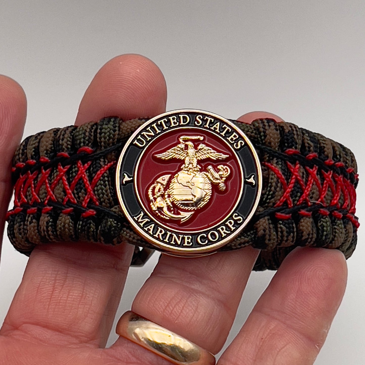 Thick, wide, rigid and comfortable braided United States Marine Corps bracelet featuring a 24K gold plated USMC Eagle Globe and Anchor medallion, a gold Cobra style tactical buckle and woven with Woodland Camo 550 paracord and Imperial Red micro and black nano cord.