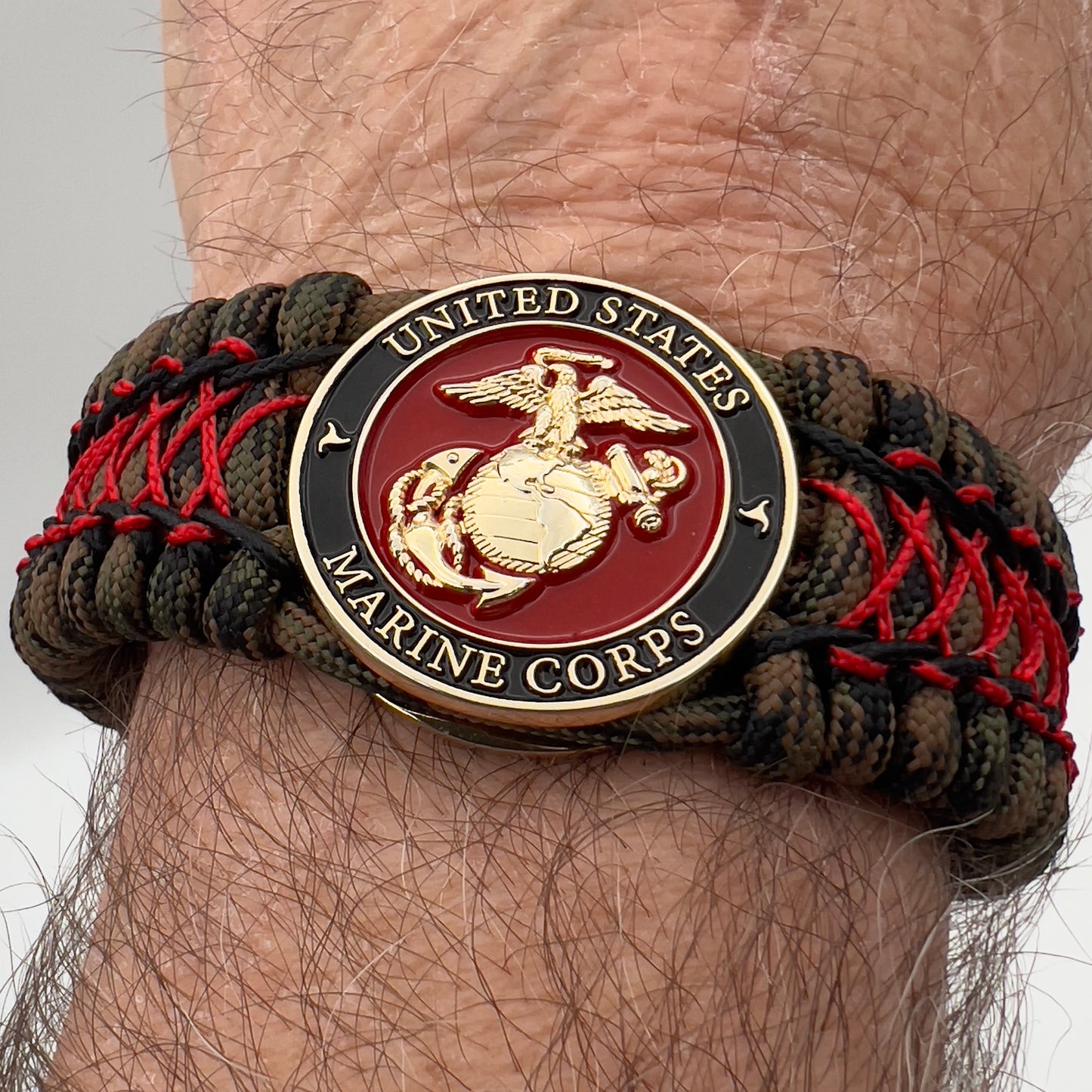 Thick, wide, rigid and comfortable braided United States Marine Corps bracelet featuring a 24K gold plated USMC Eagle Globe and Anchor medallion, a gold Cobra style tactical buckle and woven with Woodland Camo 550 paracord and Imperial Red micro and black nano cord.
