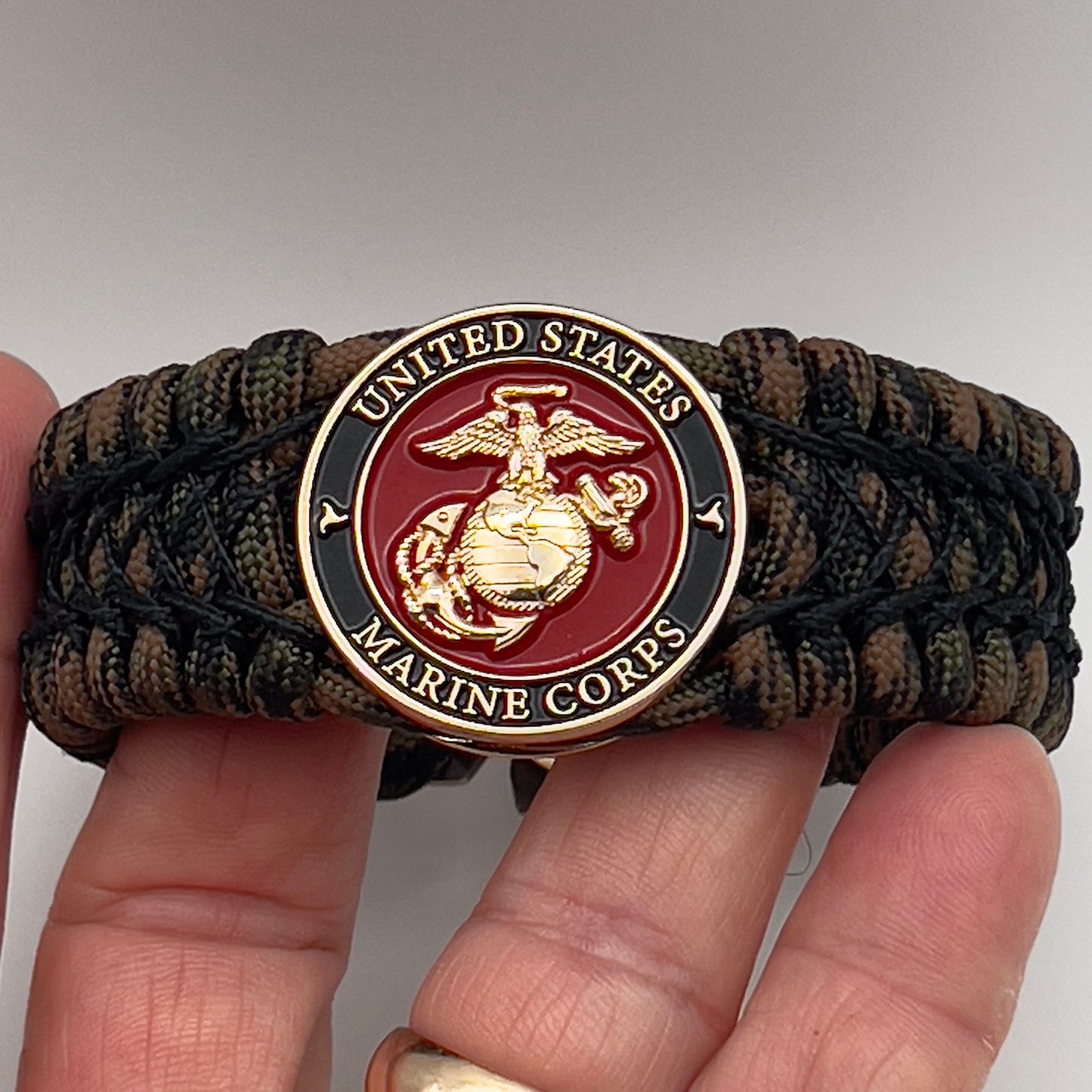 Thick, wide, rigid and comfortable braided United States Marine Corps bracelet featuring a 24K gold plated USMC Eagle Globe and Anchor medallion, a gold Cobra style tactical buckle and woven with Woodland Camo 550 paracord and black nano and micro cord.