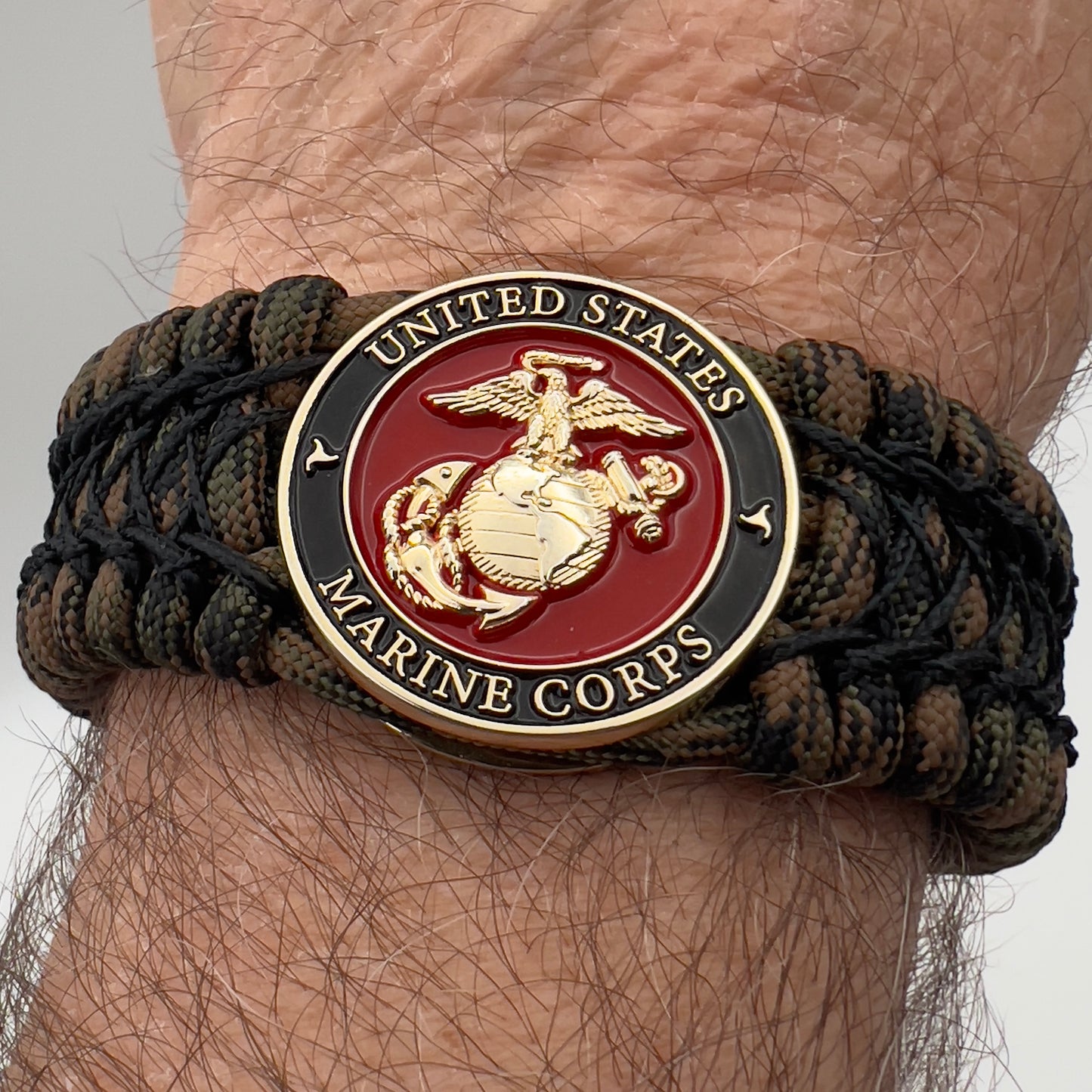 Thick, wide, rigid and comfortable braided United States Marine Corps bracelet featuring a 24K gold plated USMC Eagle Globe and Anchor medallion, a gold Cobra style tactical buckle and woven with Woodland Camo 550 paracord and black nano and micro cord.