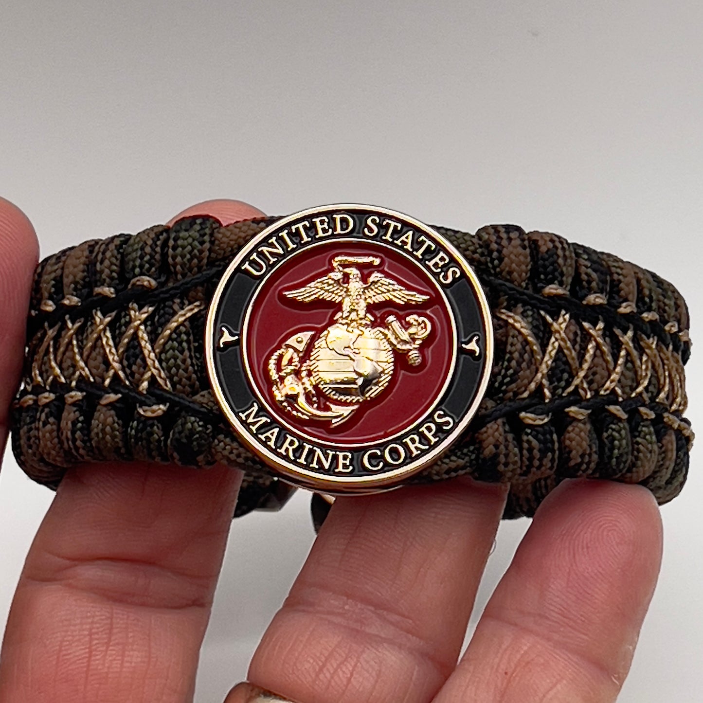 Thick, wide, rigid and comfortable braided United States Marine Corps bracelet featuring a 24K gold plated USMC Eagle Globe and Anchor medallion, a gold Cobra style tactical buckle and woven with Woodland 550 paracord, black micro and tan nano cord.