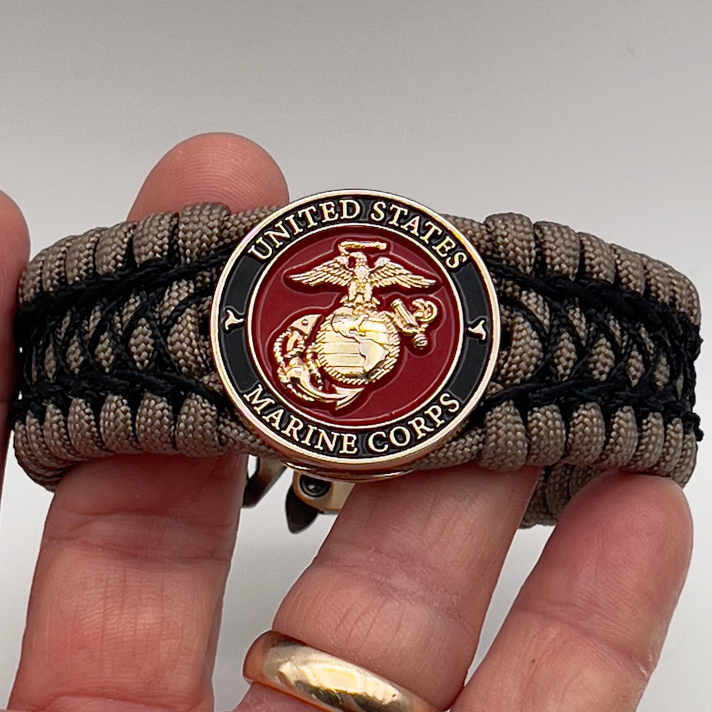 Thick, wide, rigid and comfortable braided United States Marine Corps bracelet featuring a 24K gold plated USMC Eagle Globe and Anchor medallion, a gold Cobra style tactical buckle and woven with tan 550 paracord and black nano and micro cord.