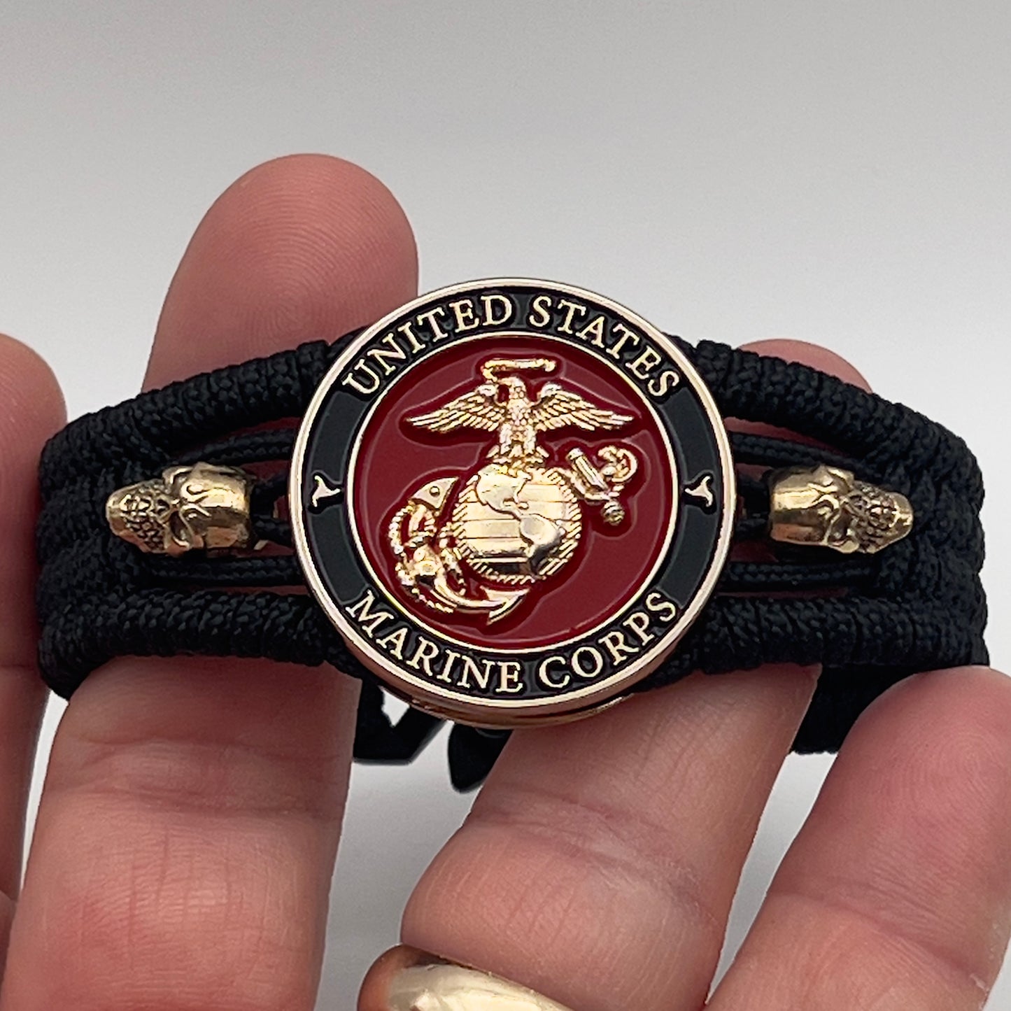 Thin, medium width, flexible and comfortable braided United States Marine Corps bracelet featuring a 24K gold plated USMC officer Eagle Globe and Anchor medallion, two gold plated skulls, a black Cobra style tactical buckle and woven with black Type-1 paracord.