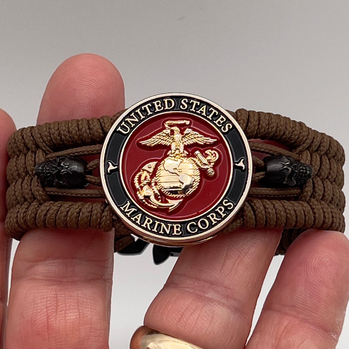Thin, medium width, flexible and comfortable braided United States Marine Corps bracelet featuring a 24K gold plated USMC officer Eagle Globe and Anchor medallion, two black micro skulls, a black Cobra style tactical buckle and woven with Coyote Brown Type-1 paracord.