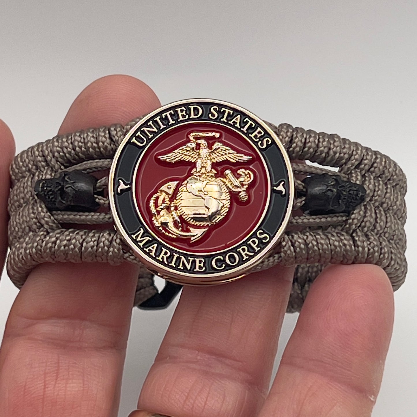 Thin, medium width, flexible and comfortable braided United States Marine Corps bracelet featuring a 24K gold plated USMC officer Eagle Globe and Anchor medallion, two black skulls, a black Cobra style tactical buckle and woven with tan Type-1 paracord.