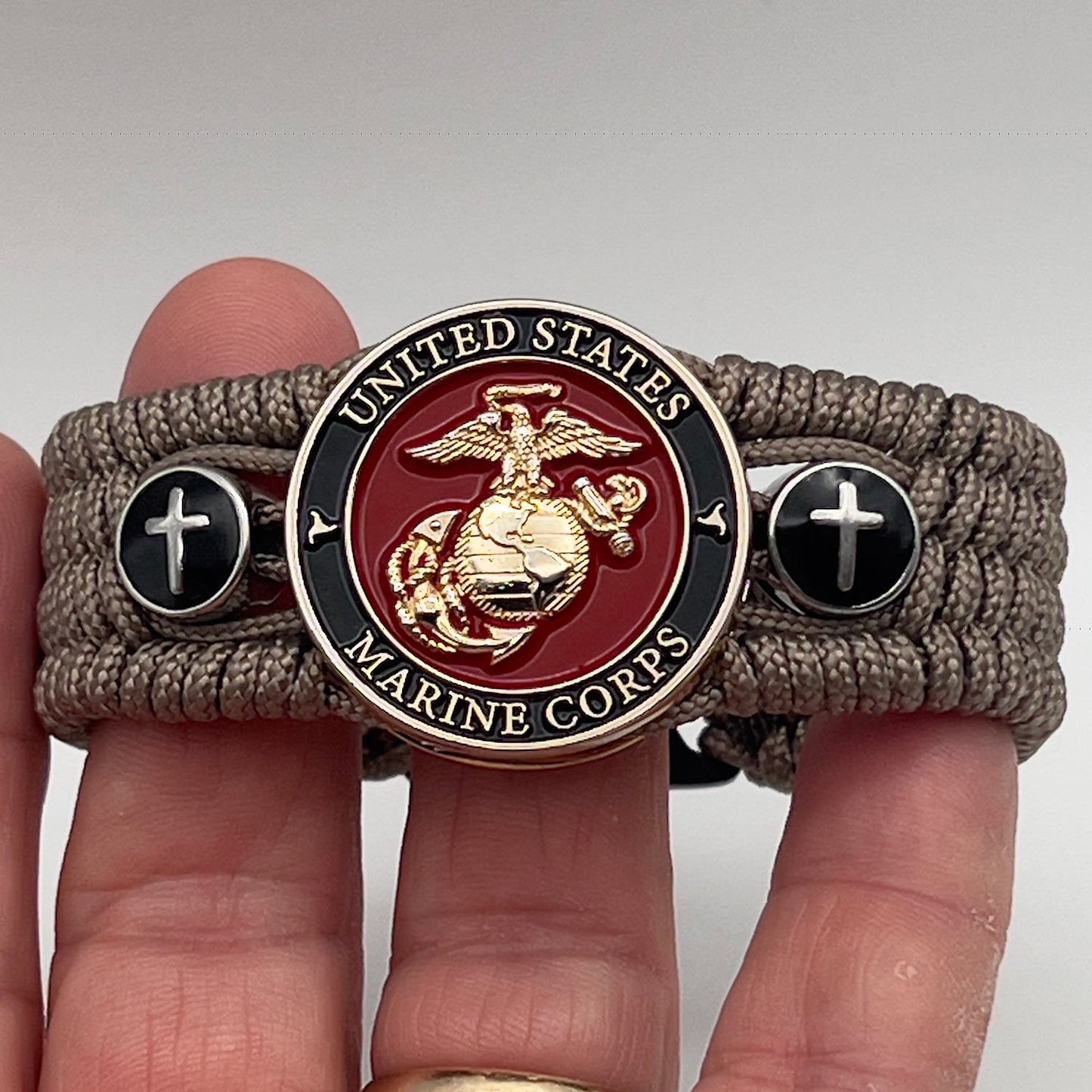 Thin, medium width, flexible and comfortable braided United States Marine Corps bracelet featuring a 24K gold plated USMC officer Eagle Globe and Anchor medallion, two Christian crosses, a black Cobra style tactical buckle and woven with Tan Type-1 paracord.