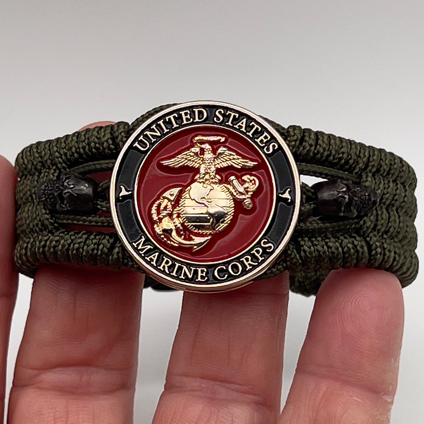 Thin, medium width, flexible and comfortable braided United States Marine Corps bracelet featuring a 24K gold plated USMC officer Eagle Globe and Anchor medallion, two black micro skulls, a black Cobra style tactical buckle and woven with Olive Drab Type-1 paracord.