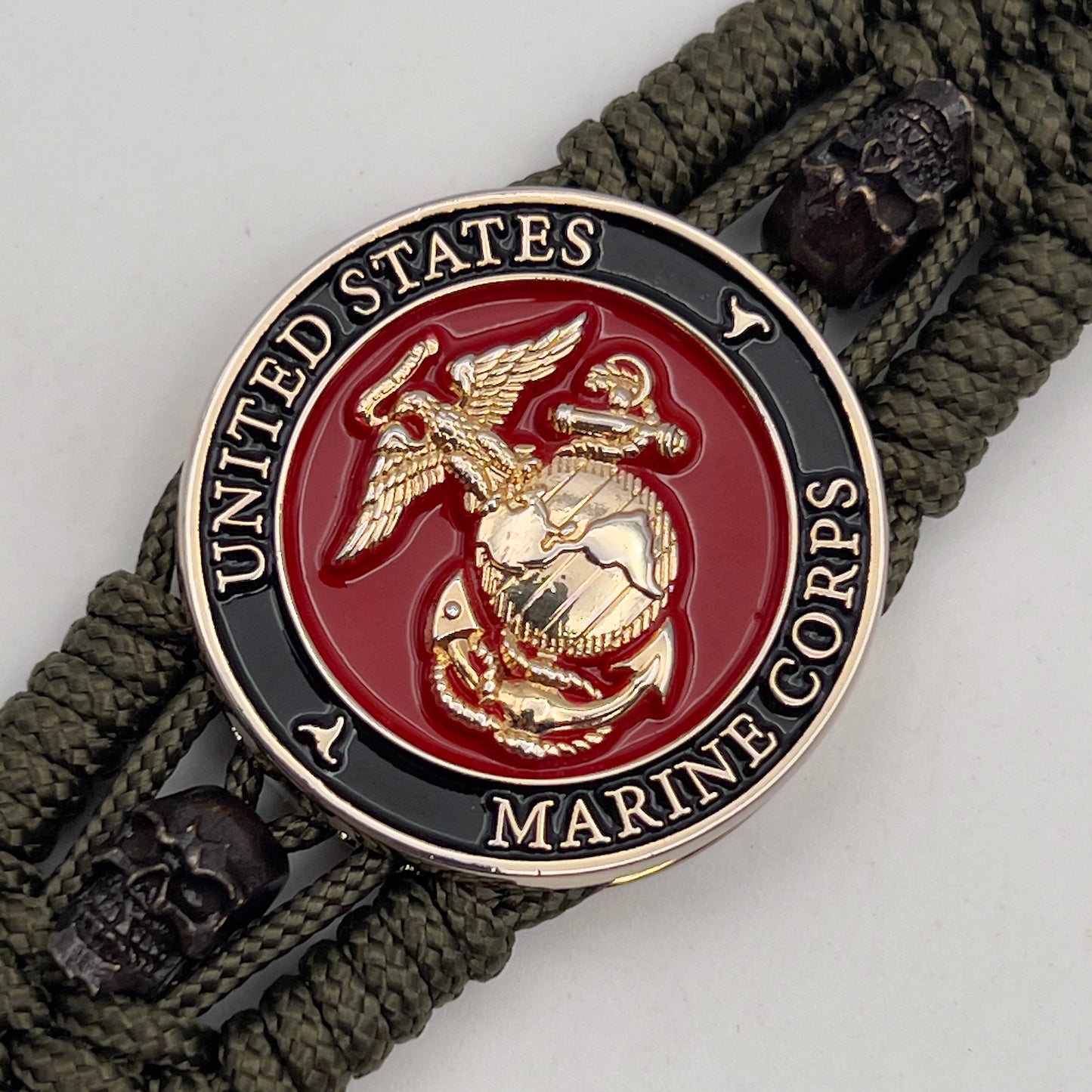 Thin, medium width, flexible and comfortable braided United States Marine Corps bracelet featuring a 24K gold plated USMC officer Eagle Globe and Anchor medallion, two black micro skulls, a black Cobra style tactical buckle and woven with Olive Drab Type-1 paracord.