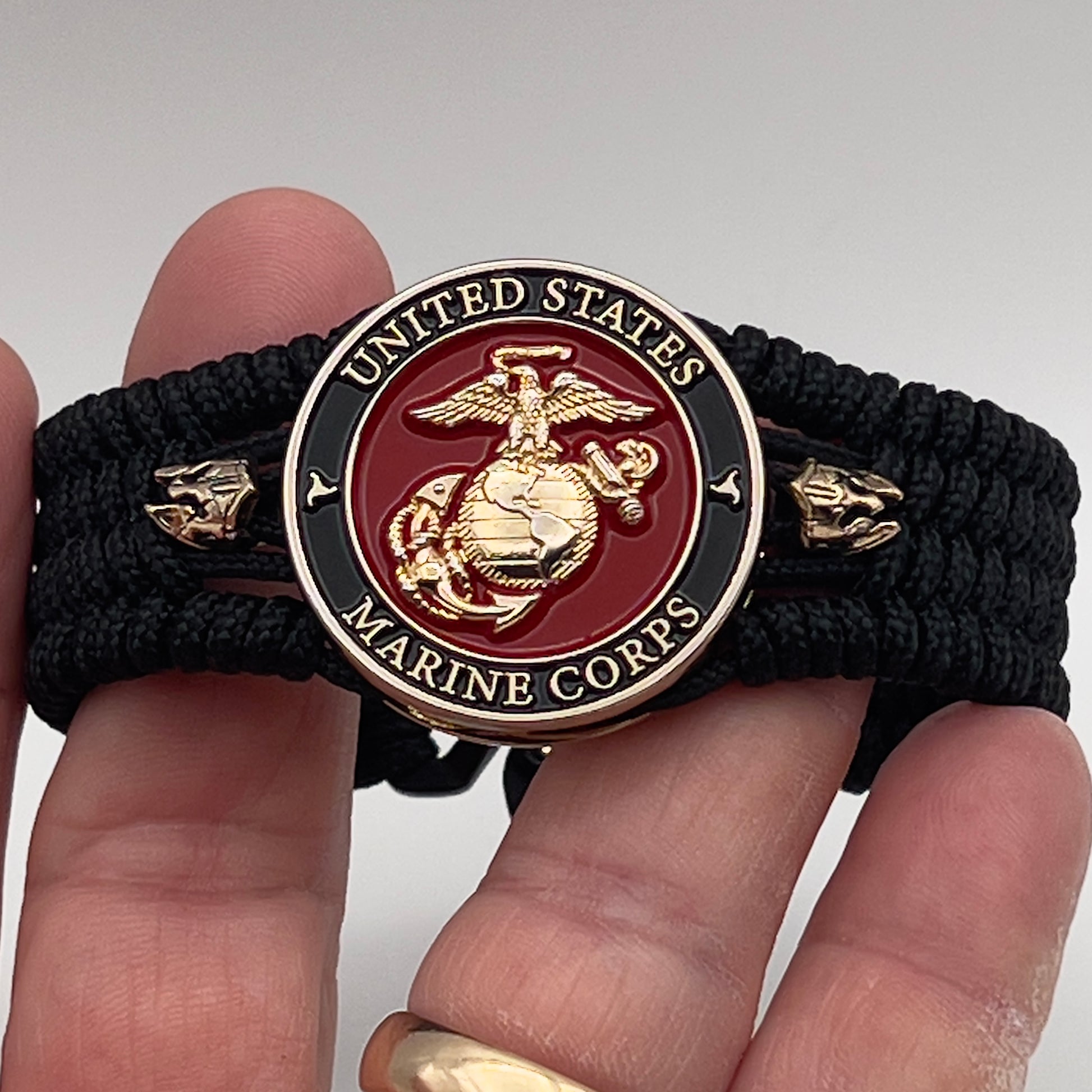 Thin, medium width, flexible and comfortable braided United States Marine Corps bracelet featuring a 24K gold plated USMC officer Eagle Globe and Anchor medallion, two brass Spartan helmets, a black Cobra style tactical buckle and woven with black Type-1 paracord.