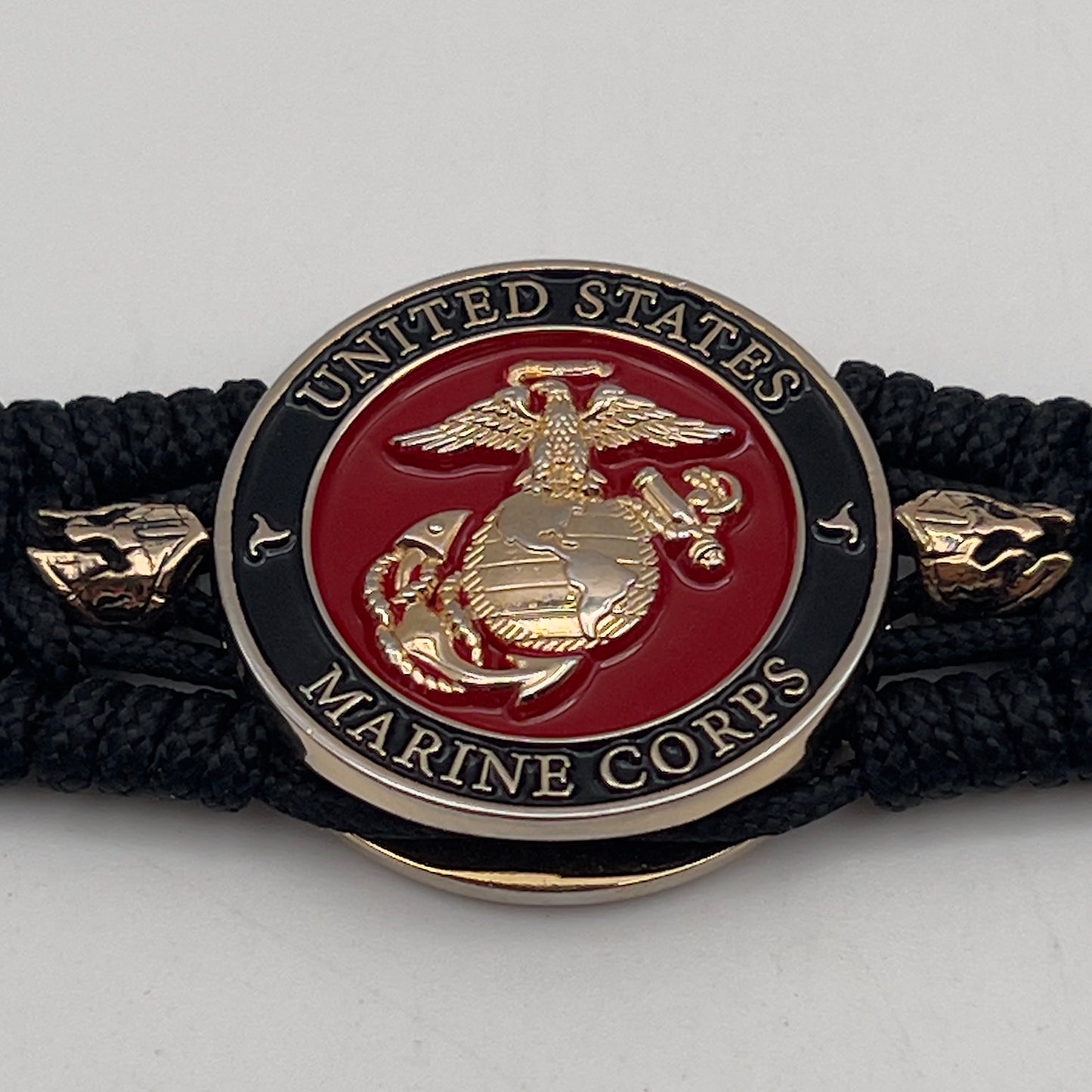 Thin, medium width, flexible and comfortable braided United States Marine Corps bracelet featuring a 24K gold plated USMC officer Eagle Globe and Anchor medallion, two brass Spartan helmets, a black Cobra style tactical buckle and woven with black Type-1 paracord.