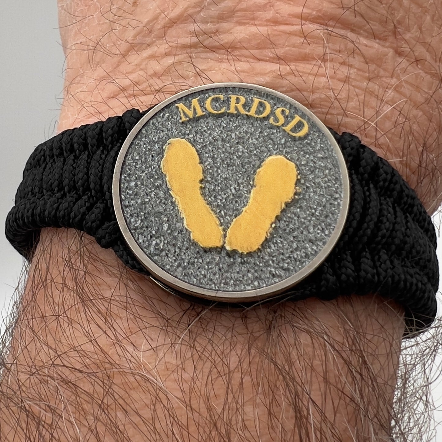 Thin, narrow, flexible and comfortable braided United States Marine Corps Recruit Depot Yellow Footprints bracelet featuring a black nickel plated UV enamel painted medallion replica of an actual set of USMC footprints, a black Cobra style tactical buckle and woven with black Type-1 paracord.