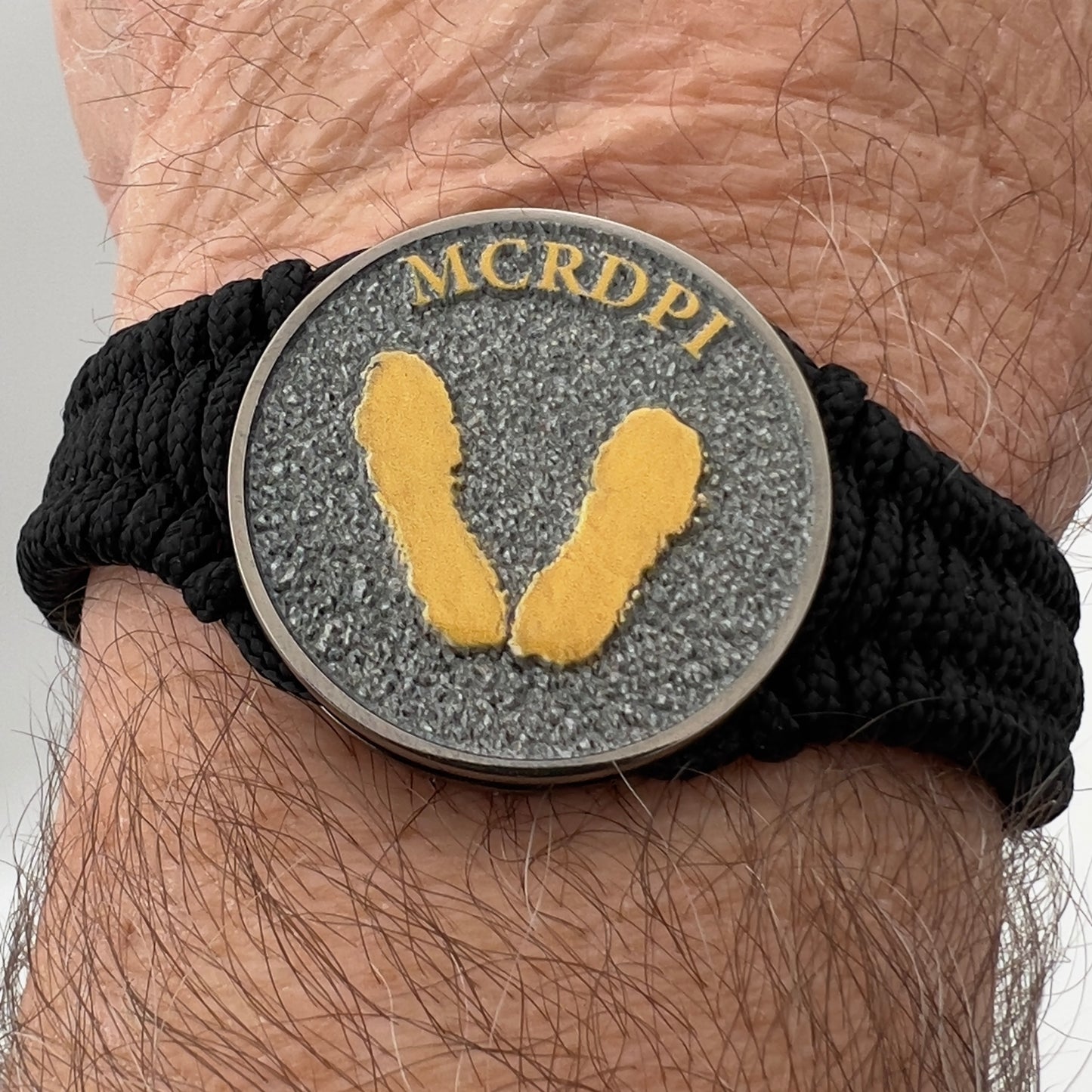 Thin, narrow, flexible and comfortable braided United States Marine Corps Recruit Depot Yellow Footprints bracelet featuring a black nickel plated UV enamel painted medallion replica of an actual set of USMC footprints, a black Cobra style tactical buckle and woven with black Type-1 paracord.