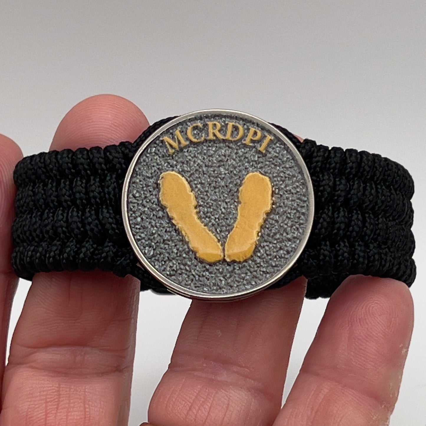 Thin, wide, flexible and comfortable braided United States Marine Corps Recruit Depot Yellow Footprints bracelet featuring a black nickel plated UV enamel painted medallion replica of an actual set of USMC footprints, a black Cobra style tactical buckle and woven with black Type-1 paracord.