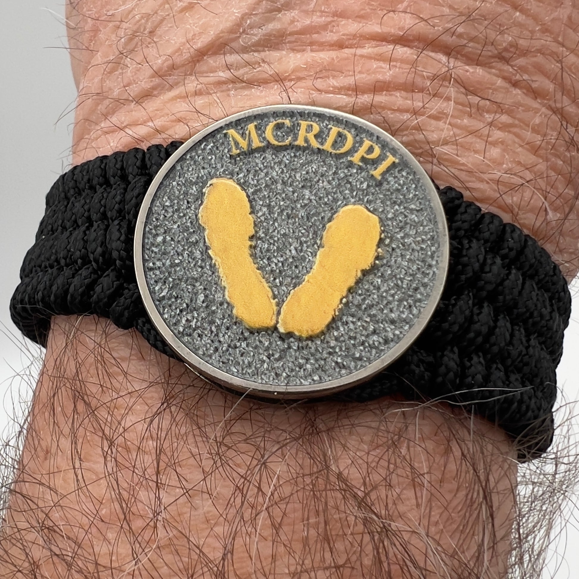 Thin, wide, flexible and comfortable braided United States Marine Corps Recruit Depot Yellow Footprints bracelet featuring a black nickel plated UV enamel painted medallion replica of an actual set of USMC footprints, a black Cobra style tactical buckle and woven with black Type-1 paracord.