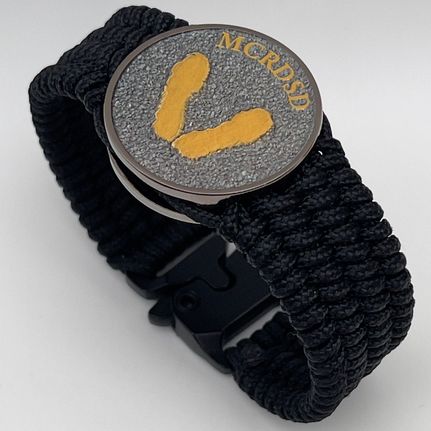 Thin, wide, flexible and comfortable braided United States Marine Corps Recruit Depot Yellow Footprints bracelet featuring a black nickel plated UV enamel painted medallion replica of an actual set of USMC footprints, a black Cobra style tactical buckle and woven with black Type-1 paracord.