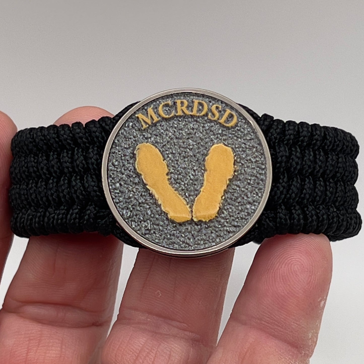 Thin, wide, flexible and comfortable braided United States Marine Corps Recruit Depot Yellow Footprints bracelet featuring a black nickel plated UV enamel painted medallion replica of an actual set of USMC footprints, a black Cobra style tactical buckle and woven with black Type-1 paracord.