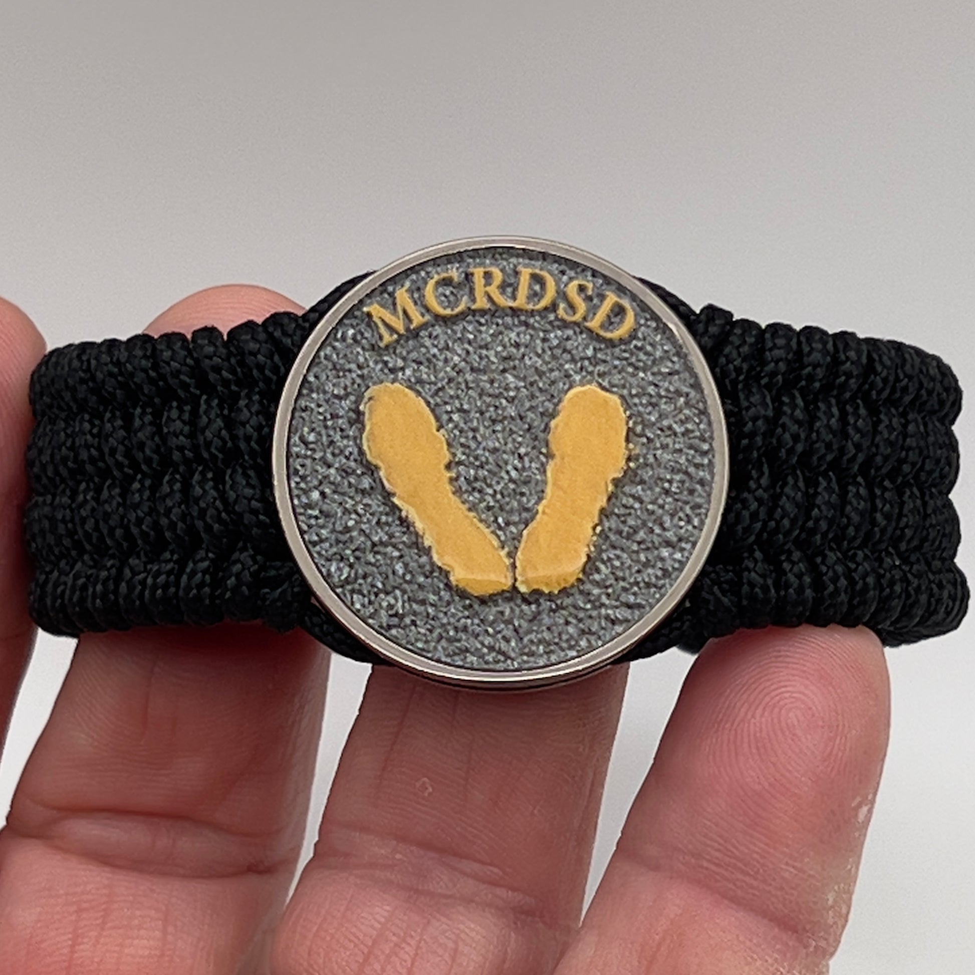 Thin, wide, flexible and comfortable braided United States Marine Corps Recruit Depot Yellow Footprints bracelet featuring a black nickel plated UV enamel painted medallion replica of an actual set of USMC footprints, a black Cobra style tactical buckle and woven with black Type-1 paracord.