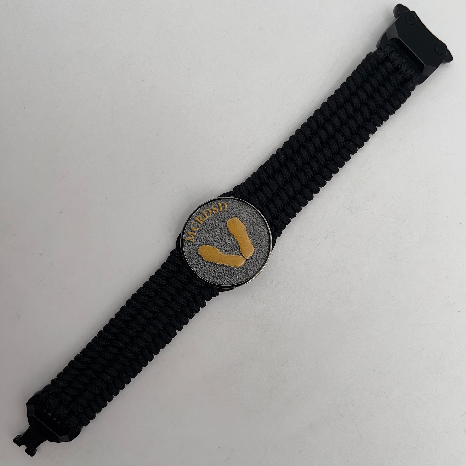 Thin, wide, flexible and comfortable braided United States Marine Corps Recruit Depot Yellow Footprints bracelet featuring a black nickel plated UV enamel painted medallion replica of an actual set of USMC footprints, a black Cobra style tactical buckle and woven with black Type-1 paracord.