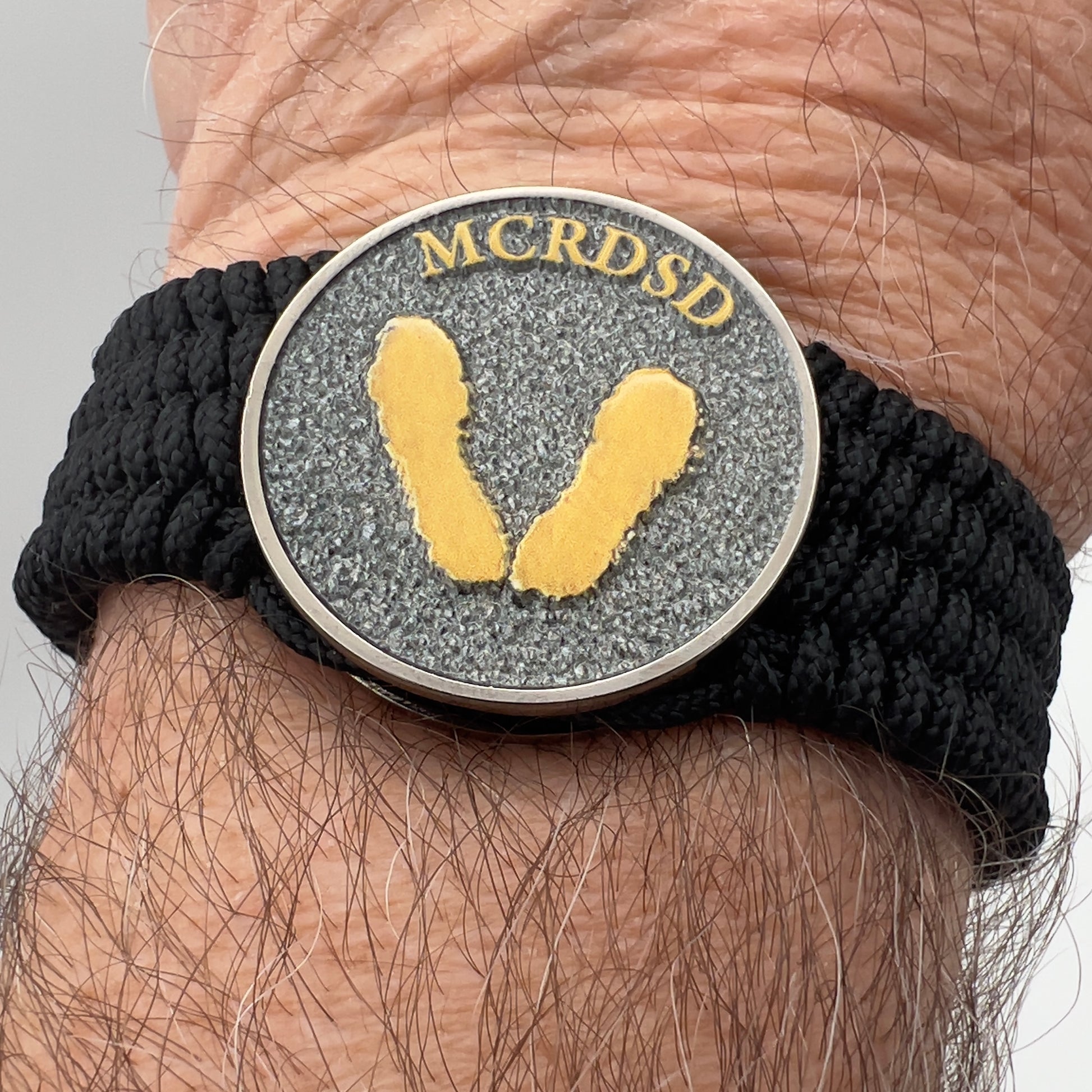 Thin, wide, flexible and comfortable braided United States Marine Corps Recruit Depot Yellow Footprints bracelet featuring a black nickel plated UV enamel painted medallion replica of an actual set of USMC footprints, a black Cobra style tactical buckle and woven with black Type-1 paracord.