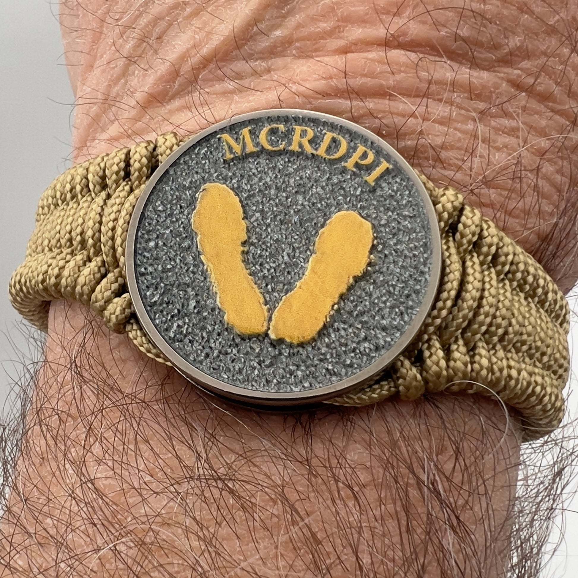 Thin, narrow, flexible and comfortable braided United States Marine Corps Recruit Depot Yellow Footprints bracelet featuring a black nickel plated UV enamel painted medallion replica of an actual set of USMC footprints, a gold Cobra style tactical buckle and woven with gold Type-1 paracord.