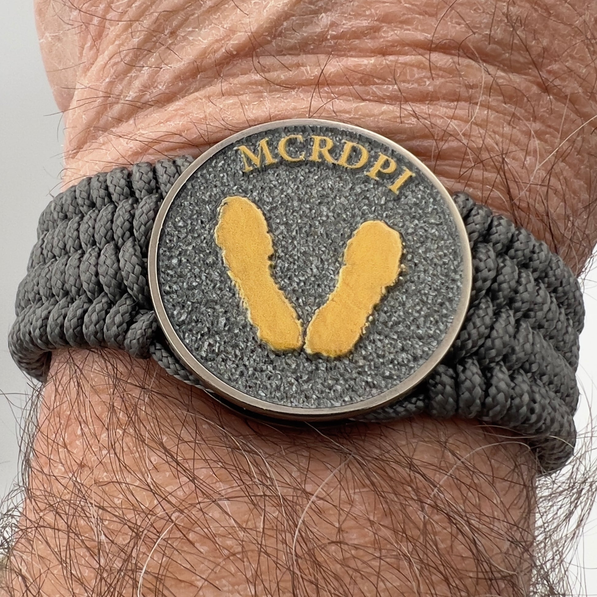 Thin, wide, flexible and comfortable braided United States Marine Corps Recruit Depot Yellow Footprints bracelet featuring a black nickel plated UV enamel painted medallion replica of an actual set of USMC footprints, a silver Cobra style tactical buckle and woven with Charcoal Gray Type-1 paracord.