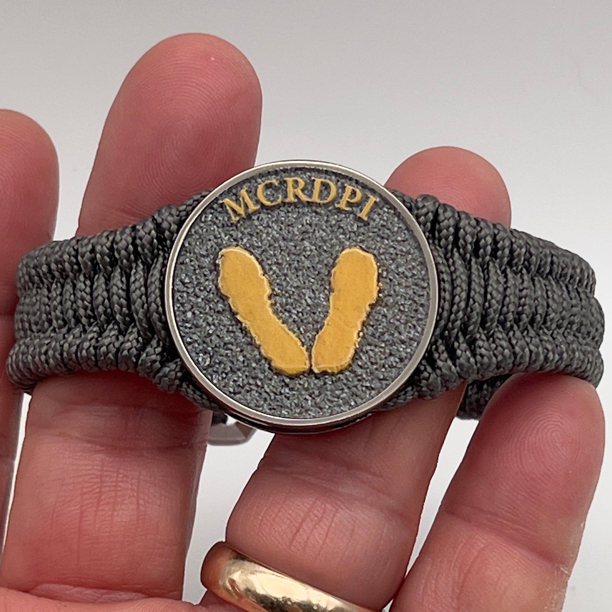 Thin, narrow, flexible and comfortable braided United States Marine Corps Recruit Depot Yellow Footprints bracelet featuring a black nickel plated UV enamel painted medallion replica of an actual set of USMC footprints, a silver Cobra style tactical buckle and woven with Charcoal Gray Type-1 paracord.