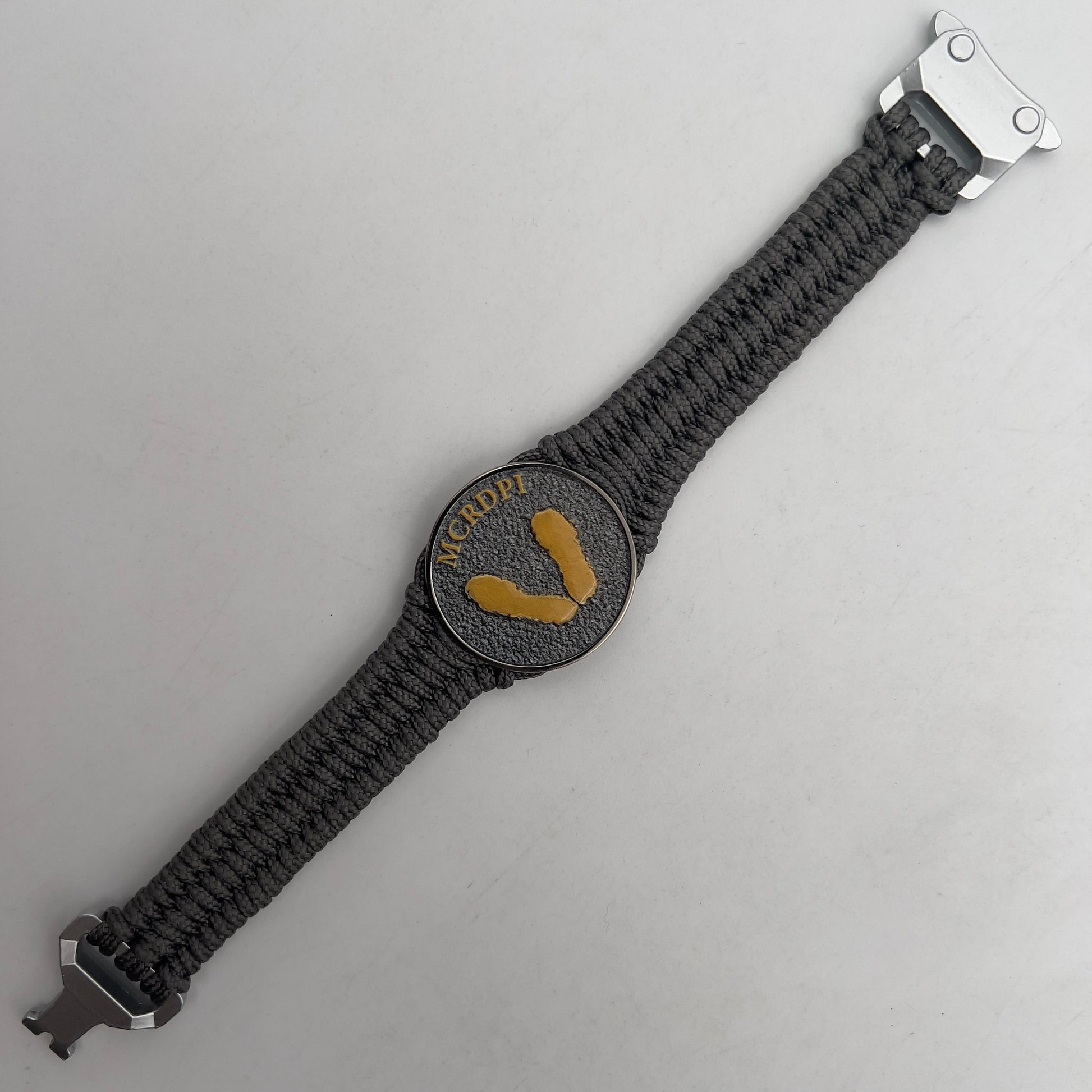Thin, narrow, flexible and comfortable braided United States Marine Corps Recruit Depot Yellow Footprints bracelet featuring a black nickel plated UV enamel painted medallion replica of an actual set of USMC footprints, a silver Cobra style tactical buckle and woven with Charcoal Gray Type-1 paracord.