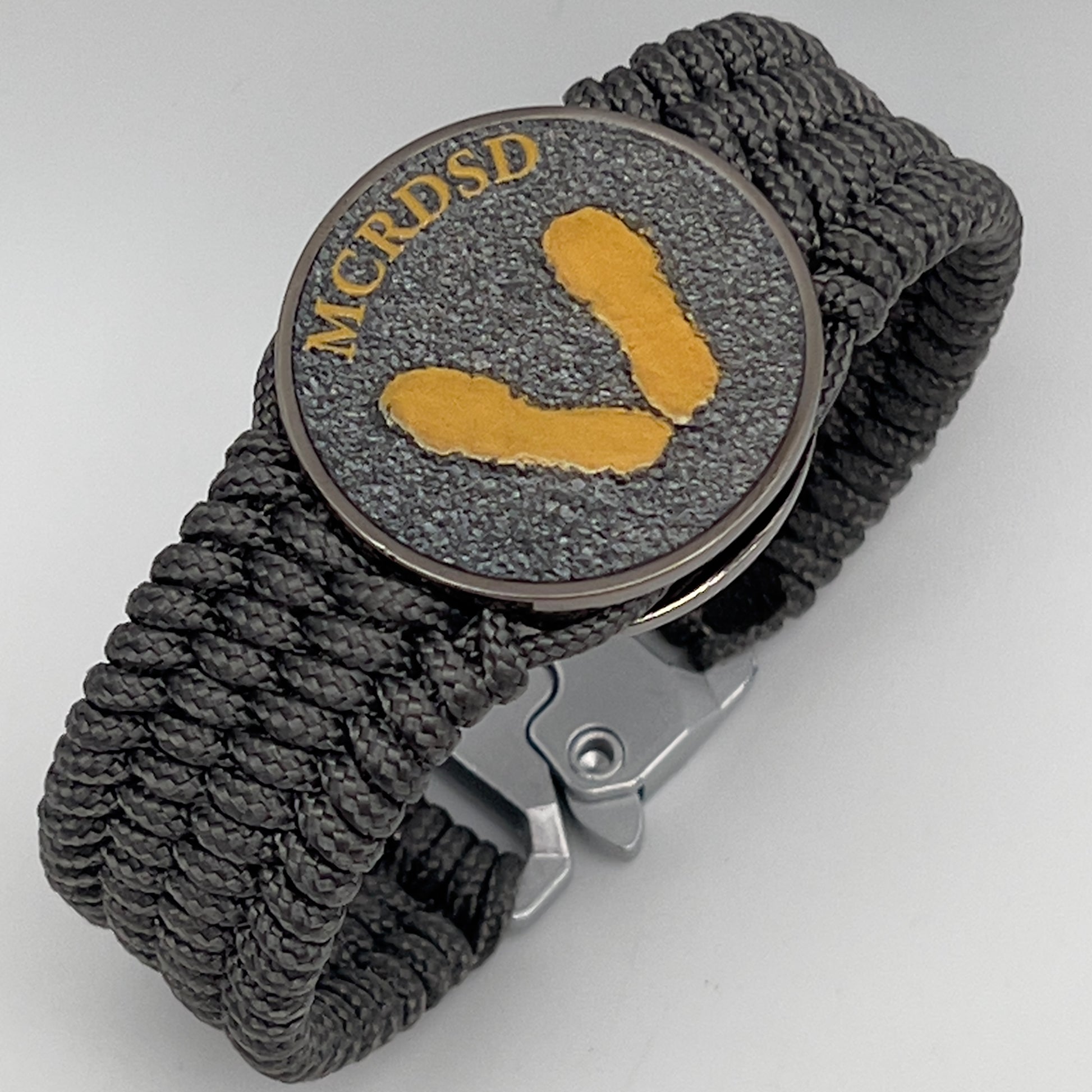 Thin, wide, flexible and comfortable braided United States Marine Corps Recruit Depot Yellow Footprints bracelet featuring a black nickel plated UV enamel painted medallion replica of an actual set of USMC footprints, a silver Cobra style tactical buckle and woven with Charcoal Gray Type-1 paracord.