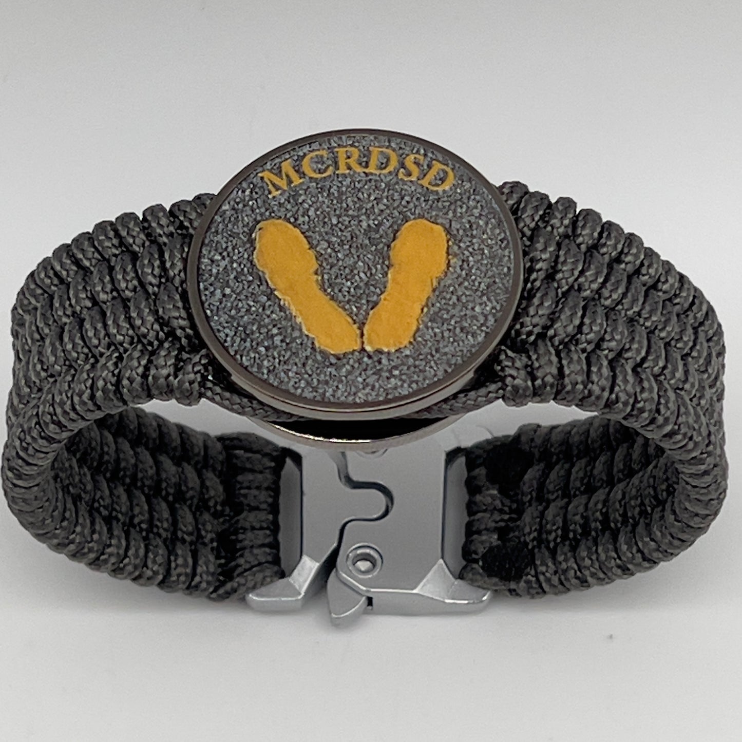 Thin, wide, flexible and comfortable braided United States Marine Corps Recruit Depot Yellow Footprints bracelet featuring a black nickel plated UV enamel painted medallion replica of an actual set of USMC footprints, a silver Cobra style tactical buckle and woven with Charcoal Gray Type-1 paracord.