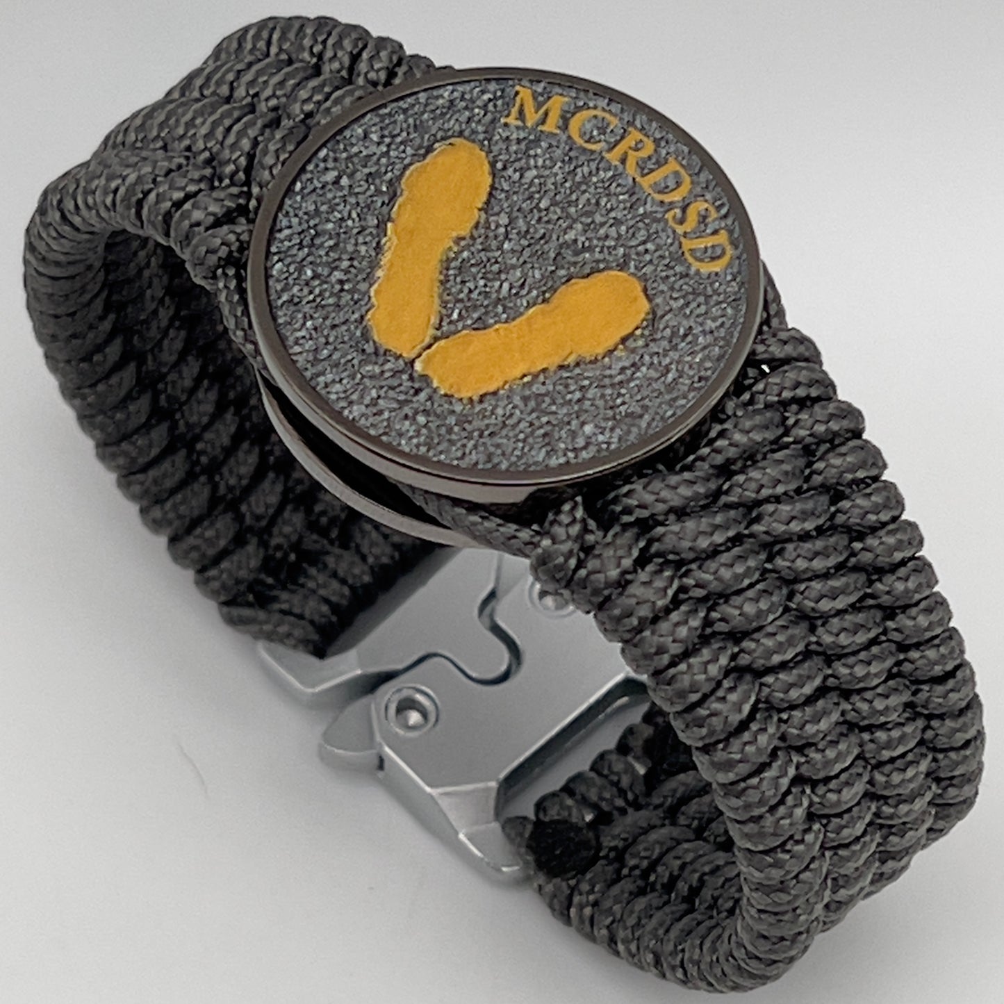Thin, wide, flexible and comfortable braided United States Marine Corps Recruit Depot Yellow Footprints bracelet featuring a black nickel plated UV enamel painted medallion replica of an actual set of USMC footprints, a silver Cobra style tactical buckle and woven with Charcoal Gray Type-1 paracord.