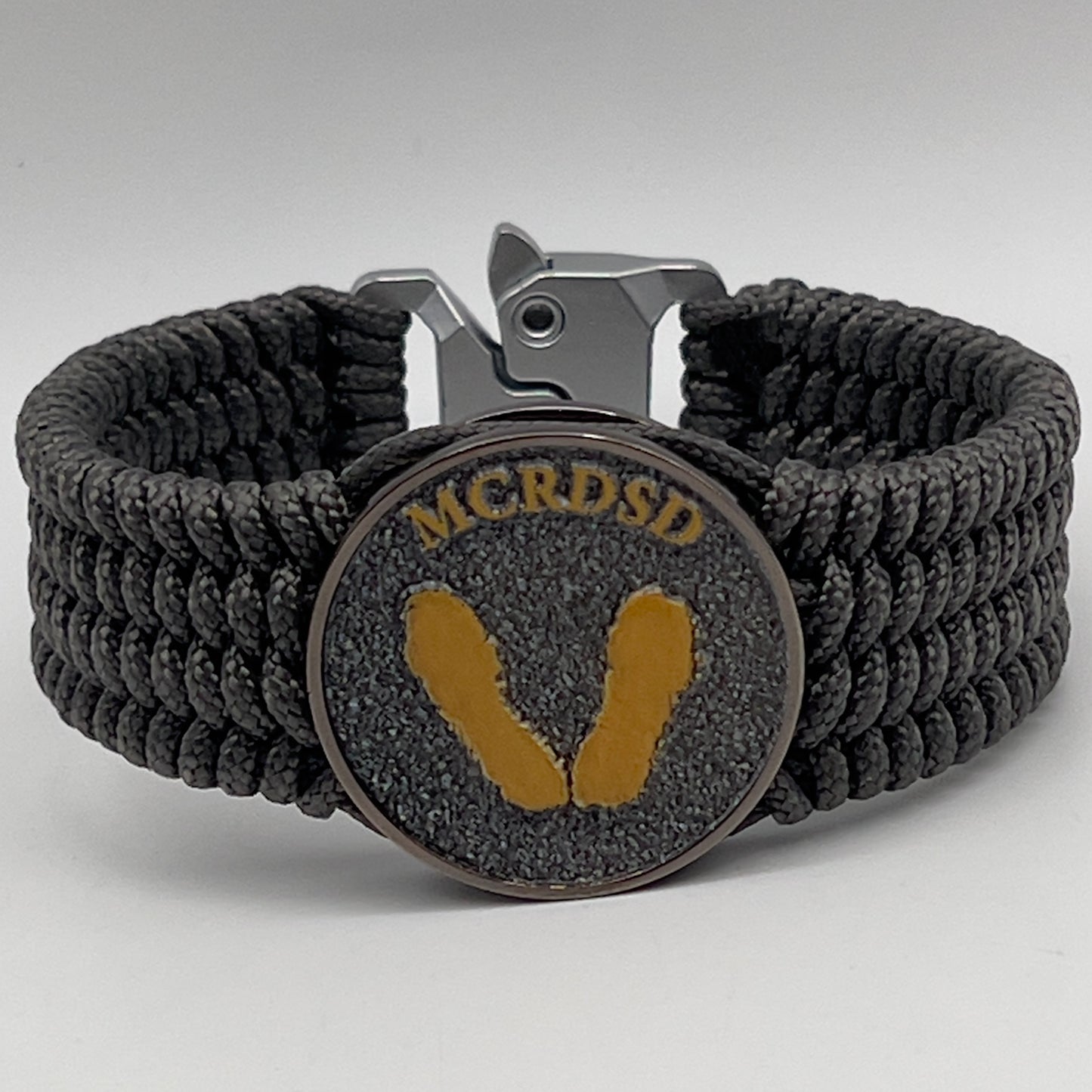 Thin, wide, flexible and comfortable braided United States Marine Corps Recruit Depot Yellow Footprints bracelet featuring a black nickel plated UV enamel painted medallion replica of an actual set of USMC footprints, a silver Cobra style tactical buckle and woven with Charcoal Gray Type-1 paracord.