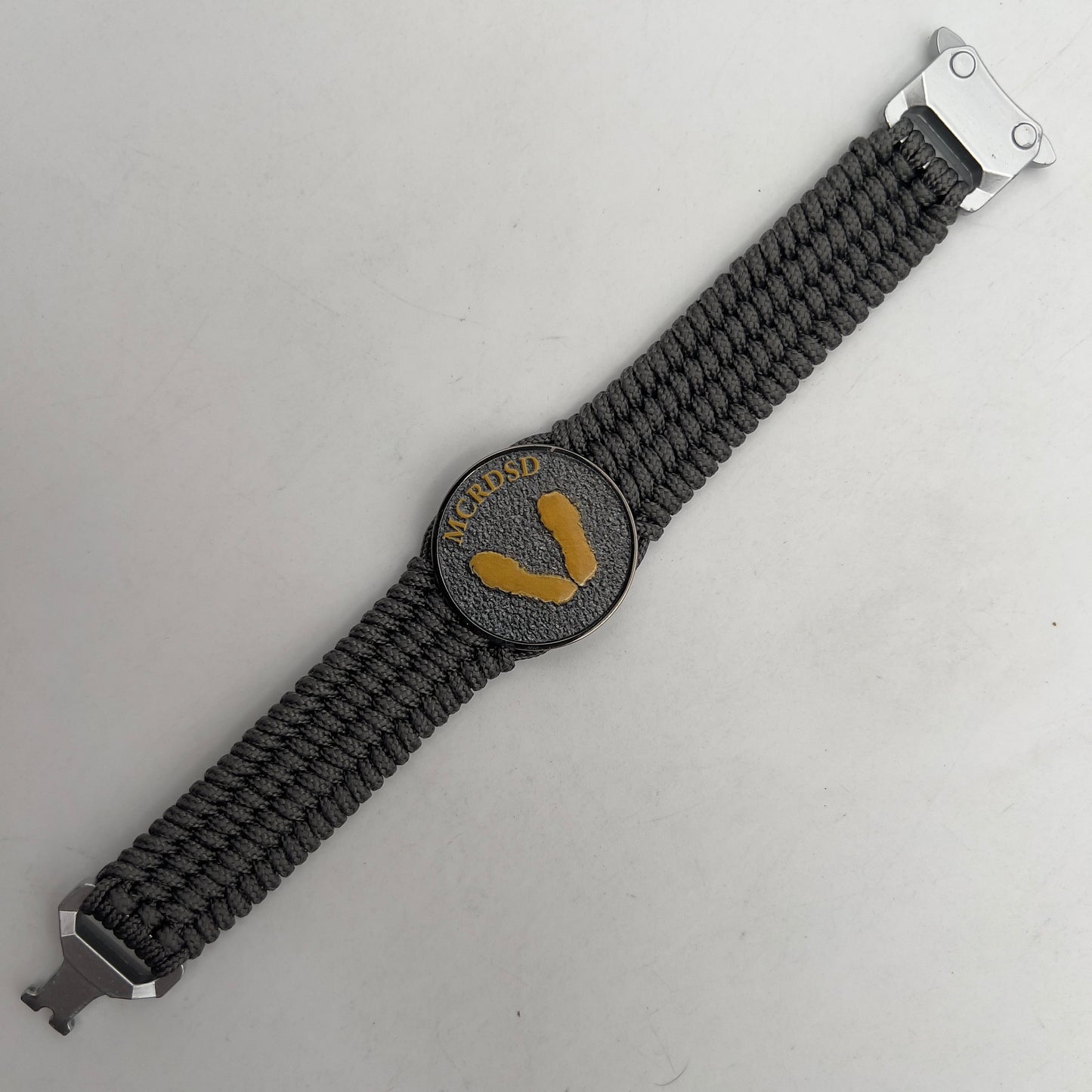 Thin, wide, flexible and comfortable braided United States Marine Corps Recruit Depot Yellow Footprints bracelet featuring a black nickel plated UV enamel painted medallion replica of an actual set of USMC footprints, a silver Cobra style tactical buckle and woven with Charcoal Gray Type-1 paracord.